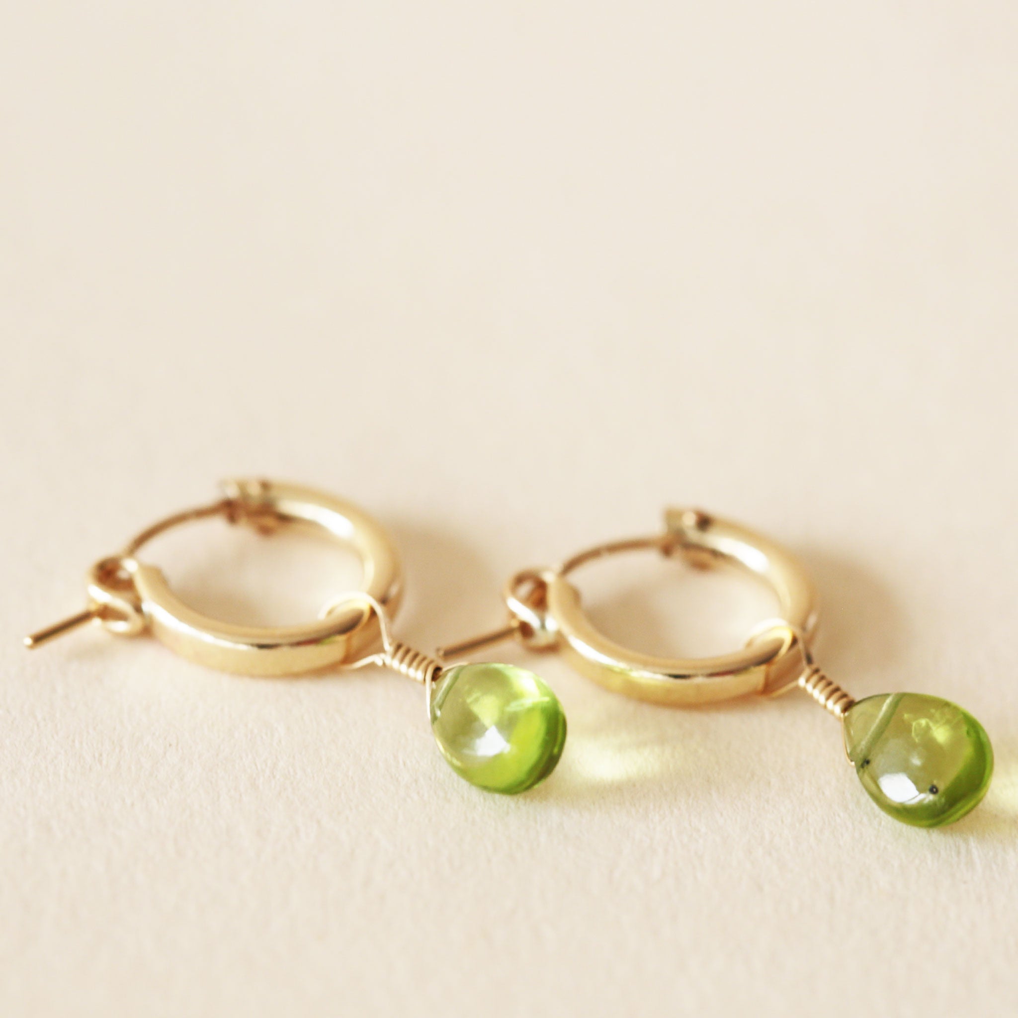 On a neutral background is a gold pair of hoop earrings with a green Peridot drop detail. 