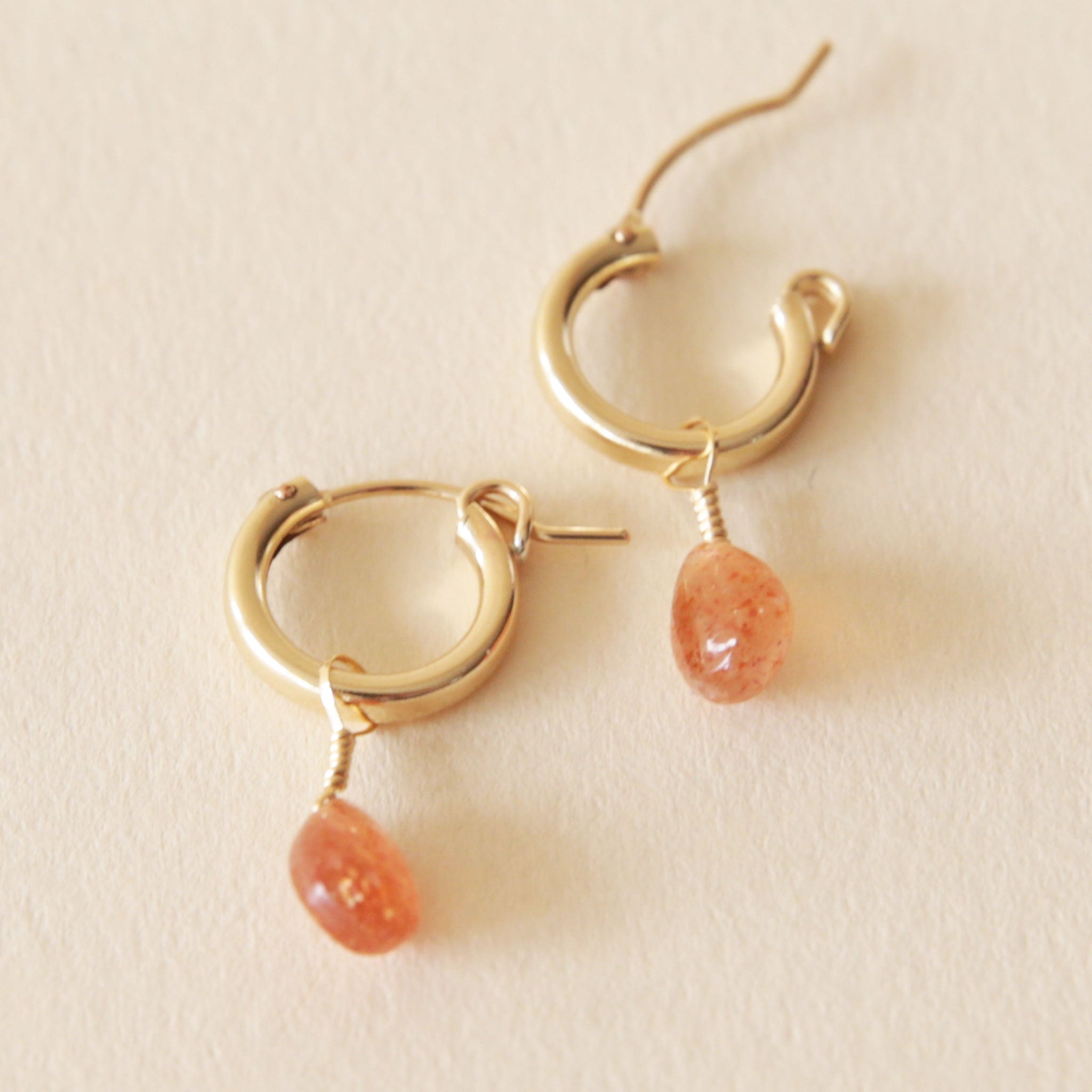 On a neutral background is a gold pair of huggie hoop earrings with a Sunstone drop gem detail. 