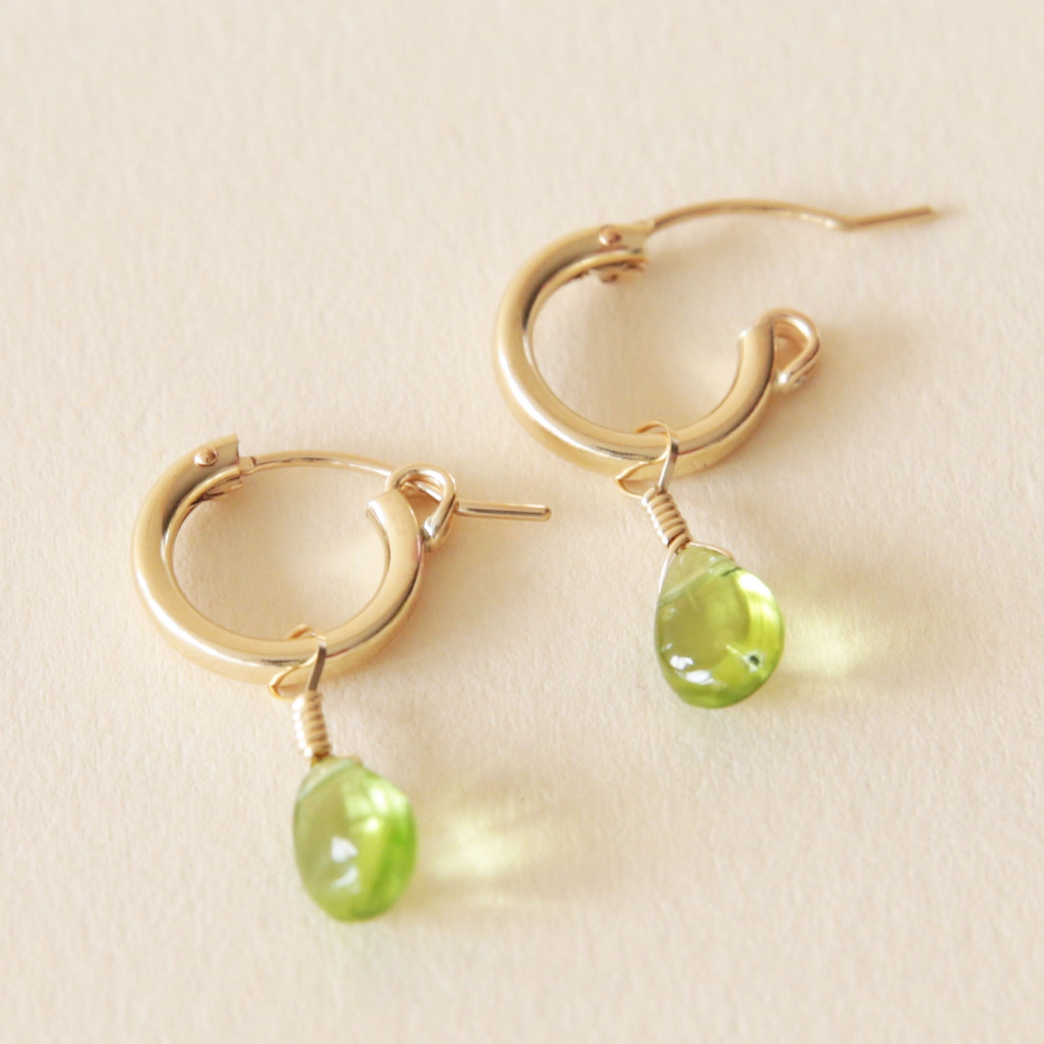 On a neutral background is a gold pair of hoop earrings with a green Peridot drop detail.