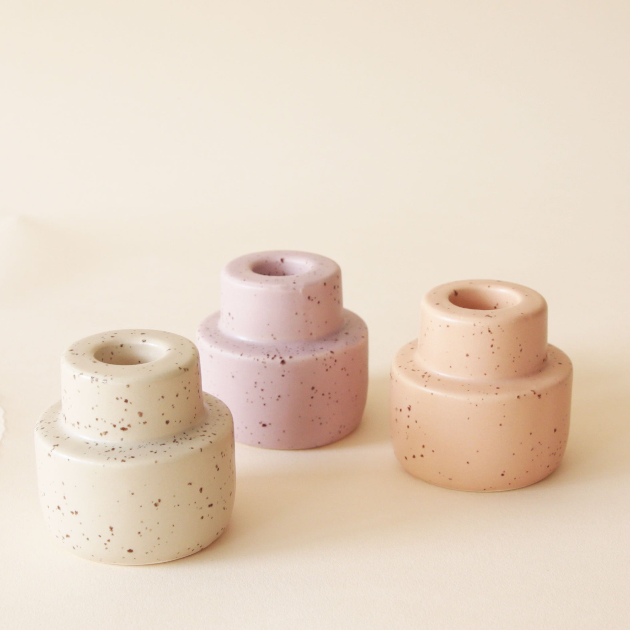 On a neutral background is three ceramic speckled candle stick holders in three different shades. From left to right there is off white, pink and peach. 