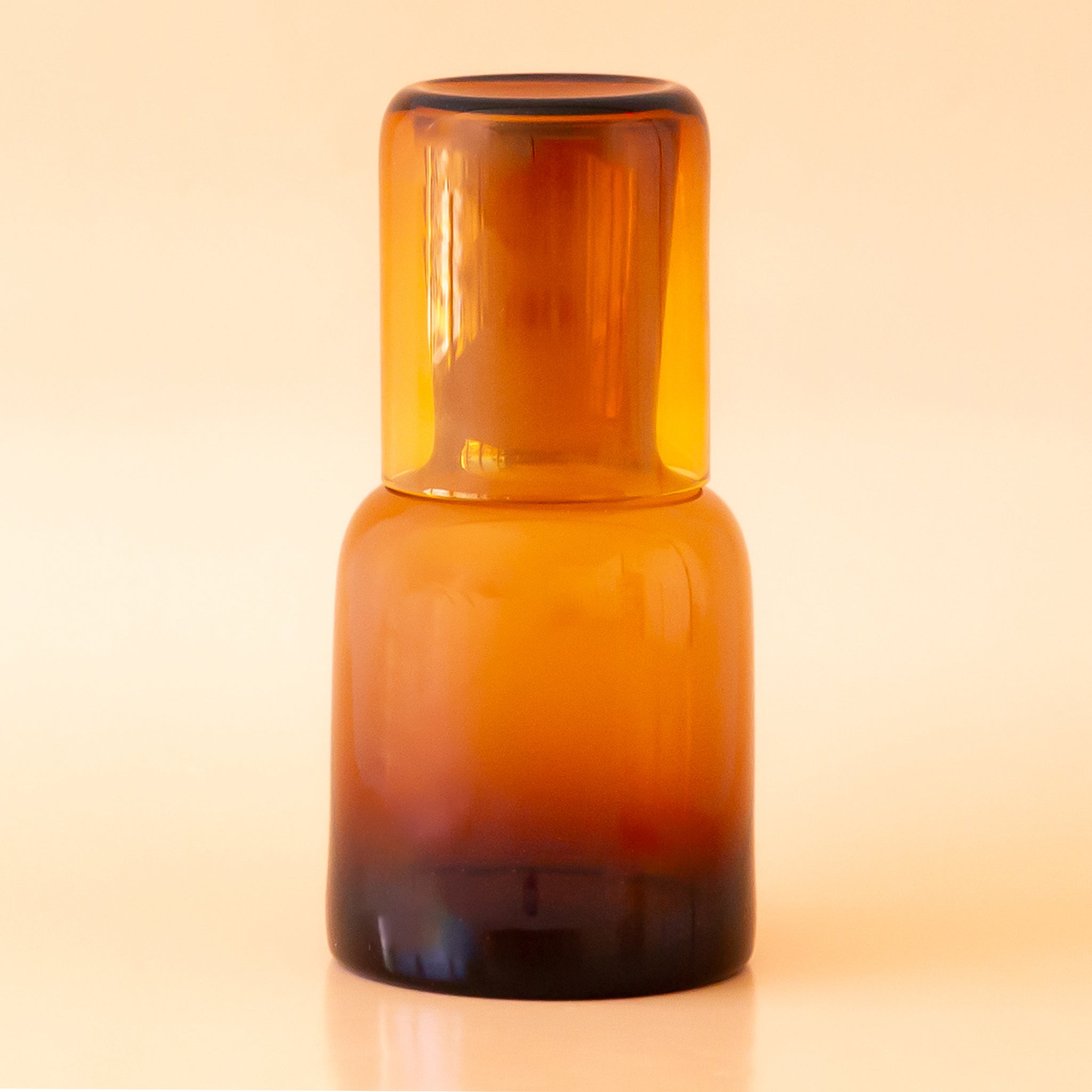 On a tan background is an amber glass carafe with a lid that doubles as a drinking glass.