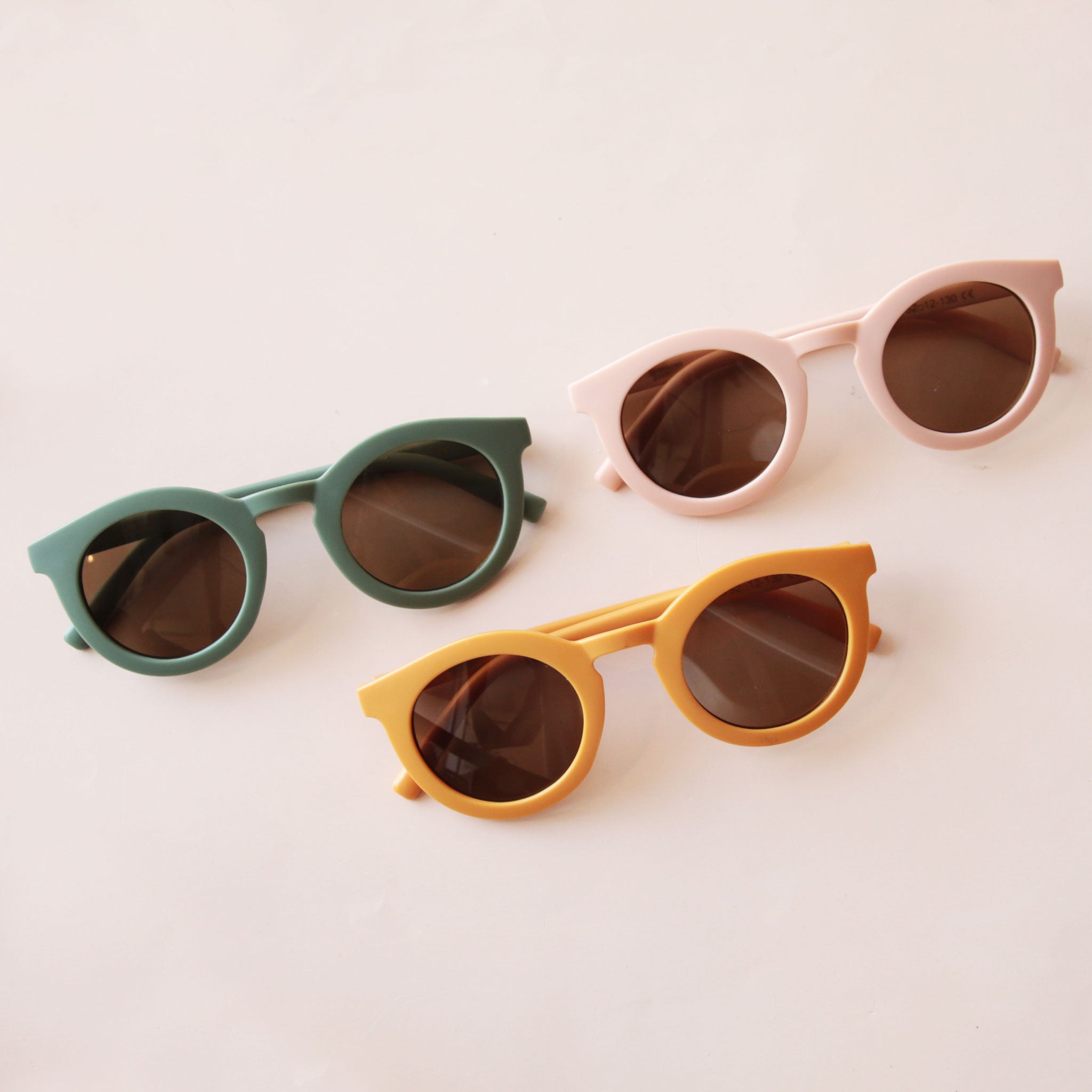 On a neutral background is three different colored circle children&#39;s sunglasses in a green, light pink and yellow shade.