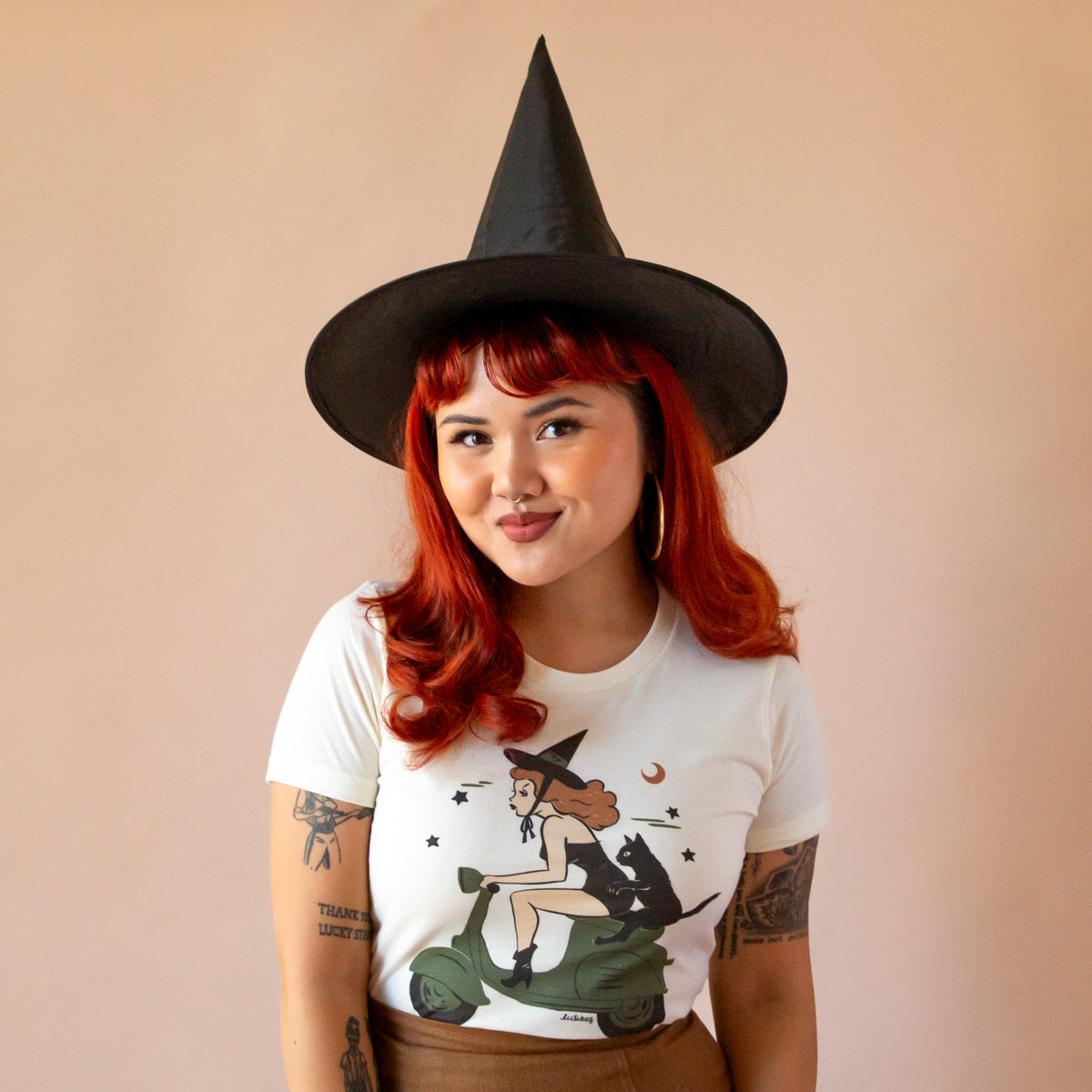 Model with red hair in ivory tee with a graphic of a witch with red hair driving a green moped with a black cat on the back.  She is wearing a witch hat. 