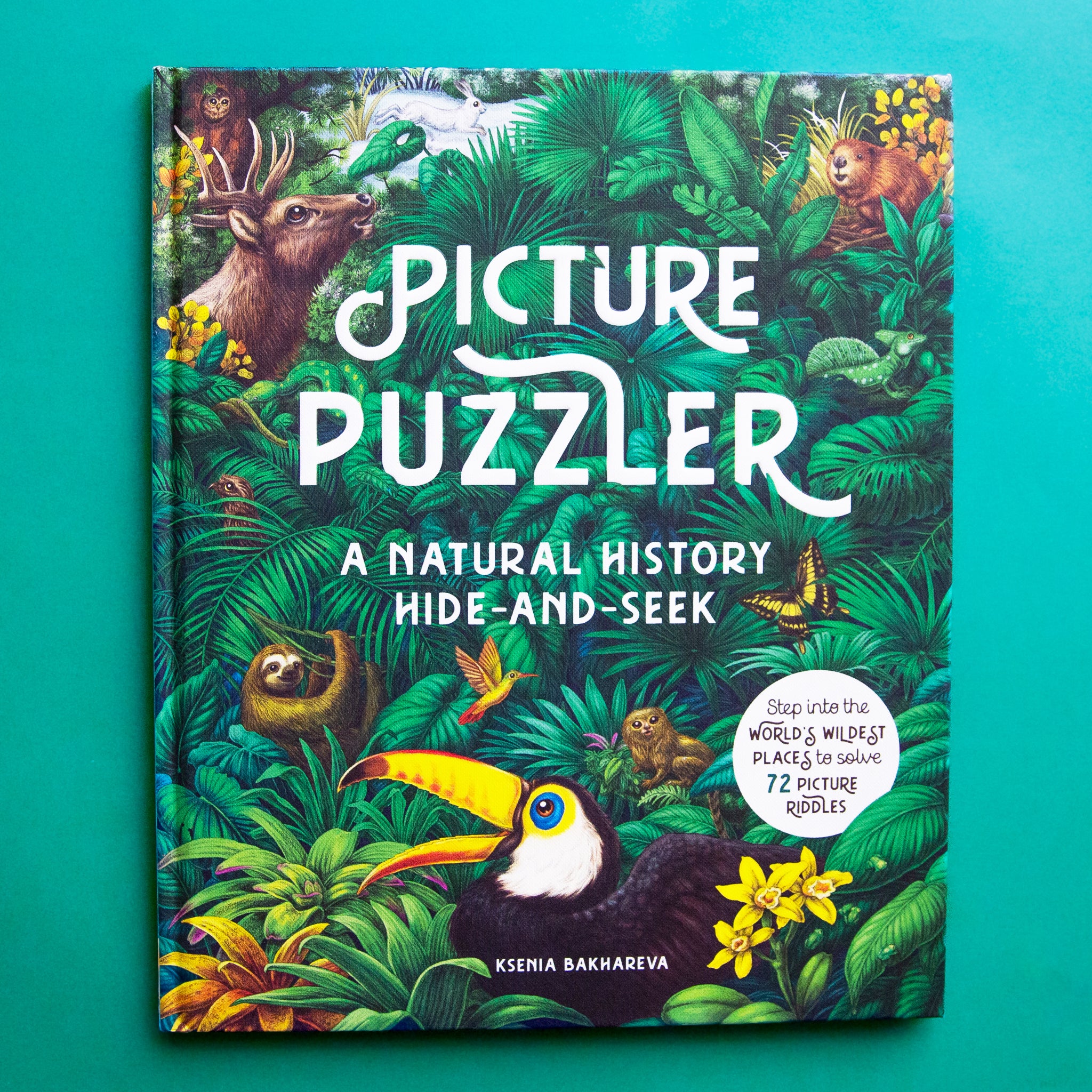 On a teal background is a book with a variety of animals and a white title in the center that reads, &quot;Picture Puzzler A Natural History Hide-And-Seek&quot;. 