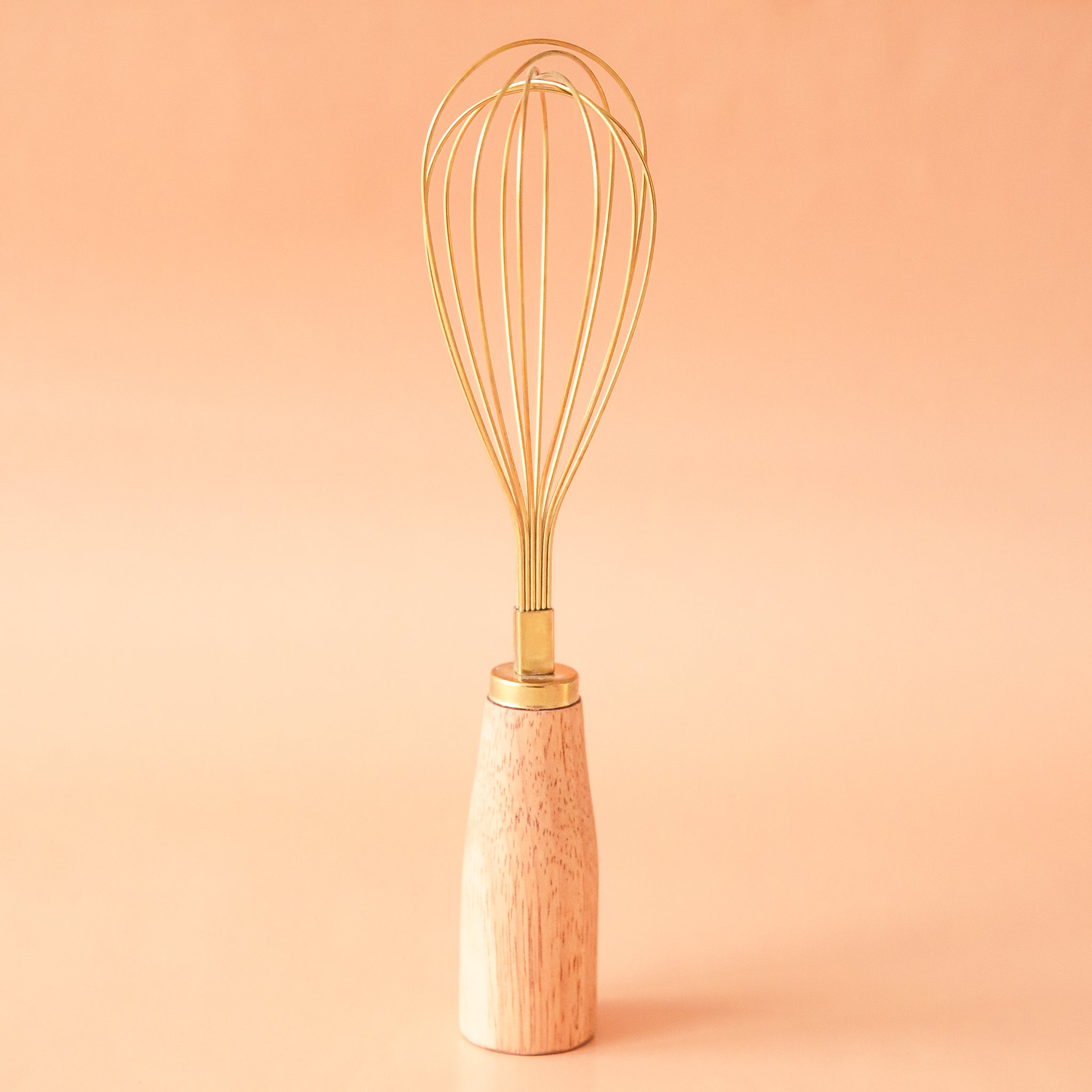 On a peachy background is a gold whisk with a wood handle.