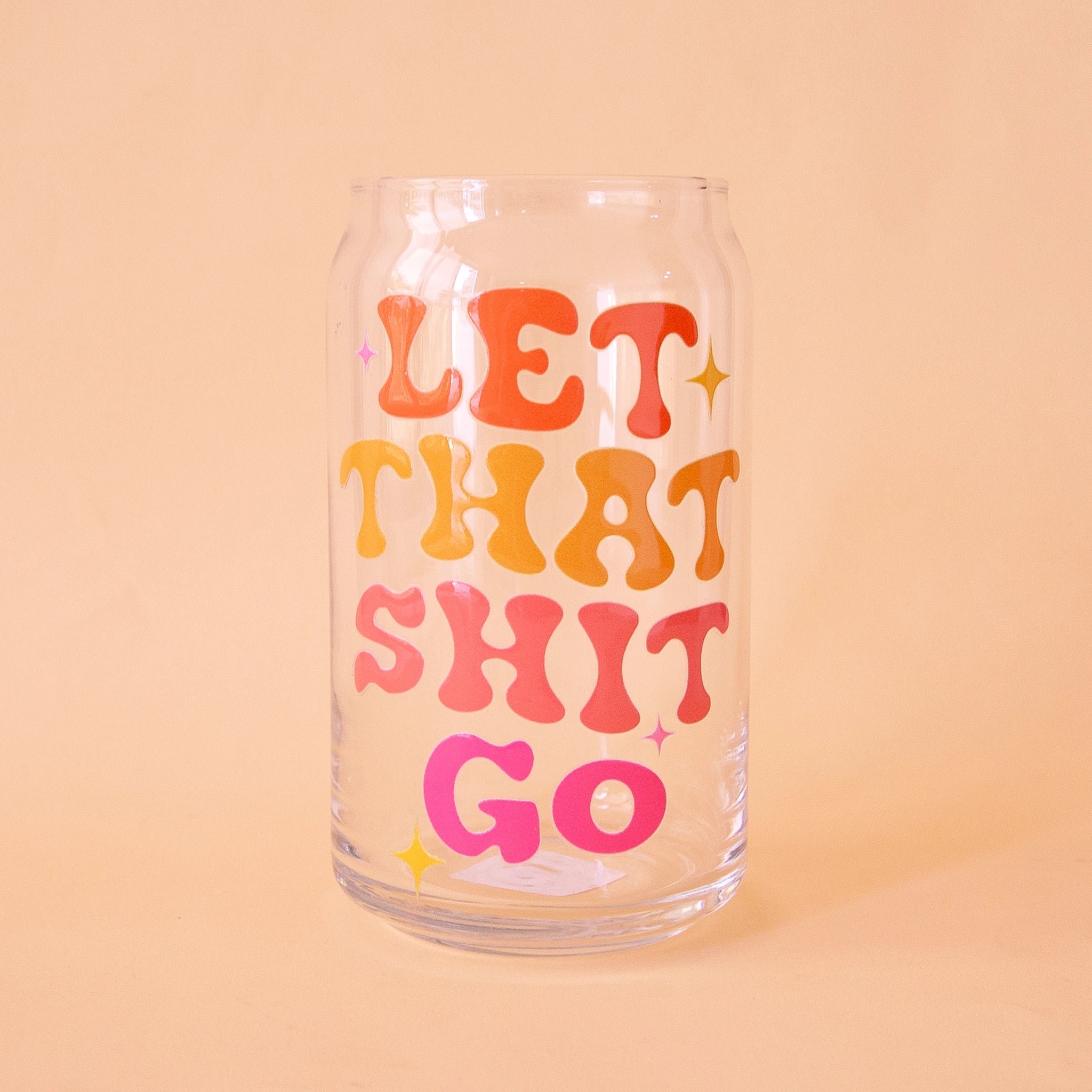 On a peach background is a drinking glass with red, pink and orange words on the front that reads, "Let That Shit Go".