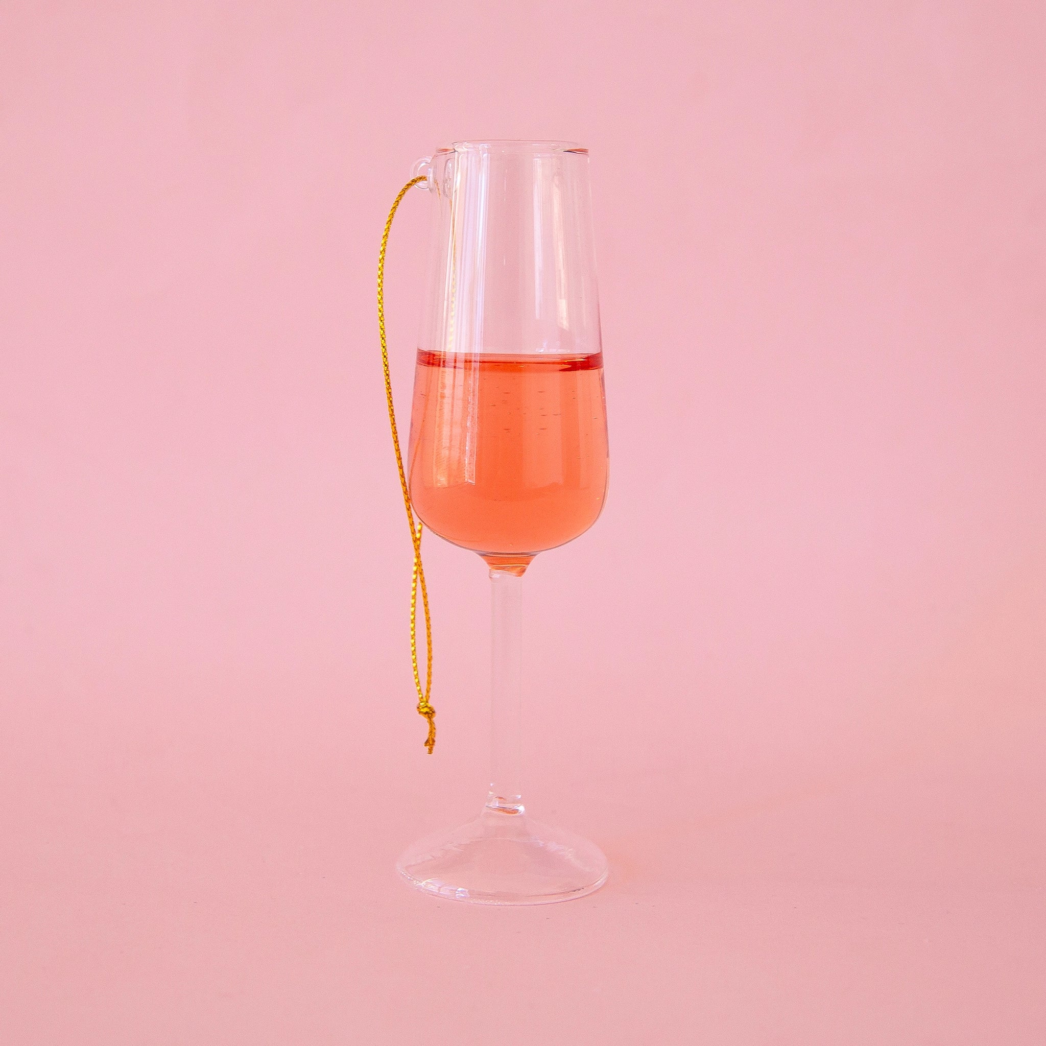 On a bright pink background is a glass ornament in the shape of a champagne flute with a pink bottom half resembling rose. 