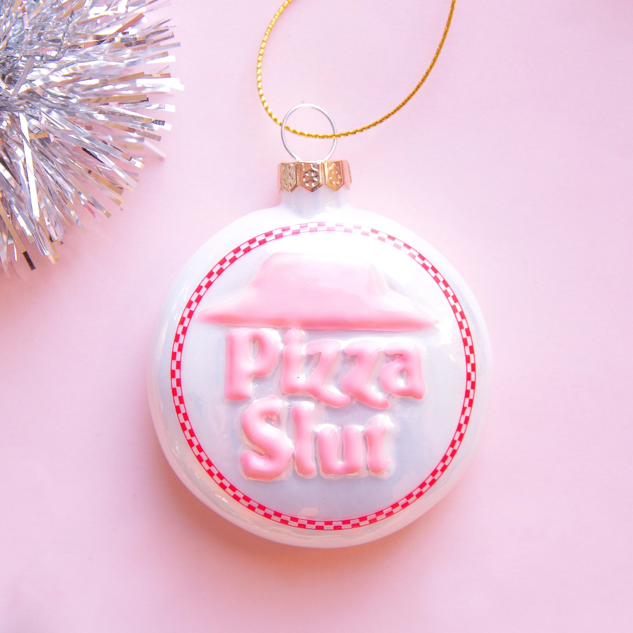 On a pink snowy background is a white and pink circle glass ornament that has text in the center that reads, &quot;Pizza Slut&quot;.