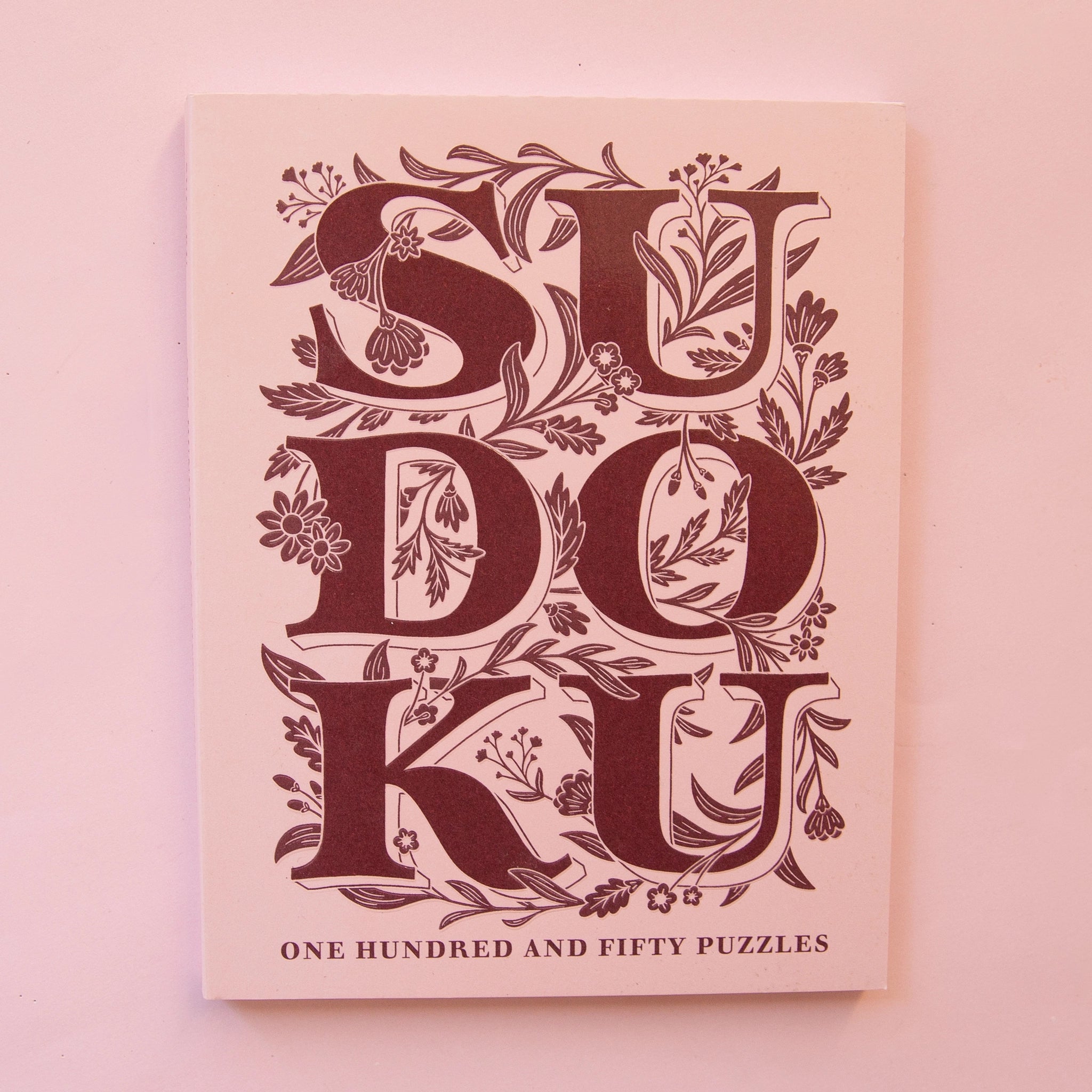 Pastel pink paperback puzzle book titled &#39;Sudoku&#39; in large magenta capital lettering. The letters are accented with delightful floral detailing. Below the large lettering reads &#39;One hundred and fifty puzzles&#39; towards the bottom of the cover.