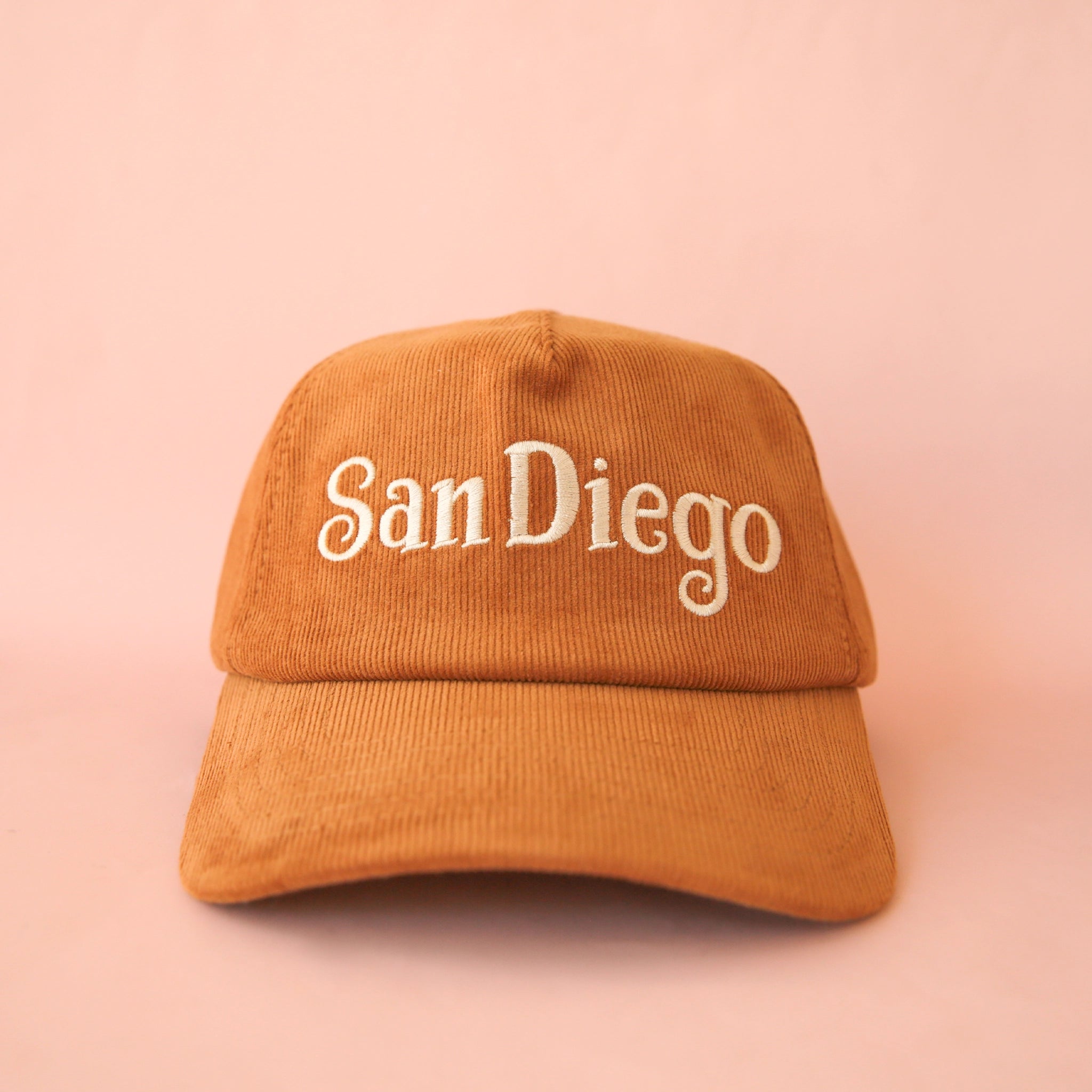 On a pink background is a toffee colored baseball cap that has a corduroy texture and says, "San Diego" in white lettering.