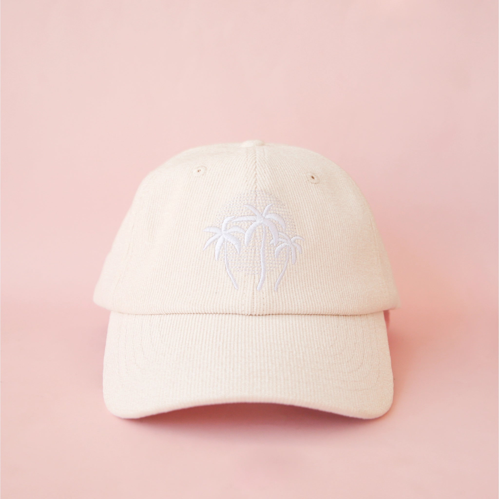 On a pink background is an ivory baseball hat with three palm tree logo in the center. 