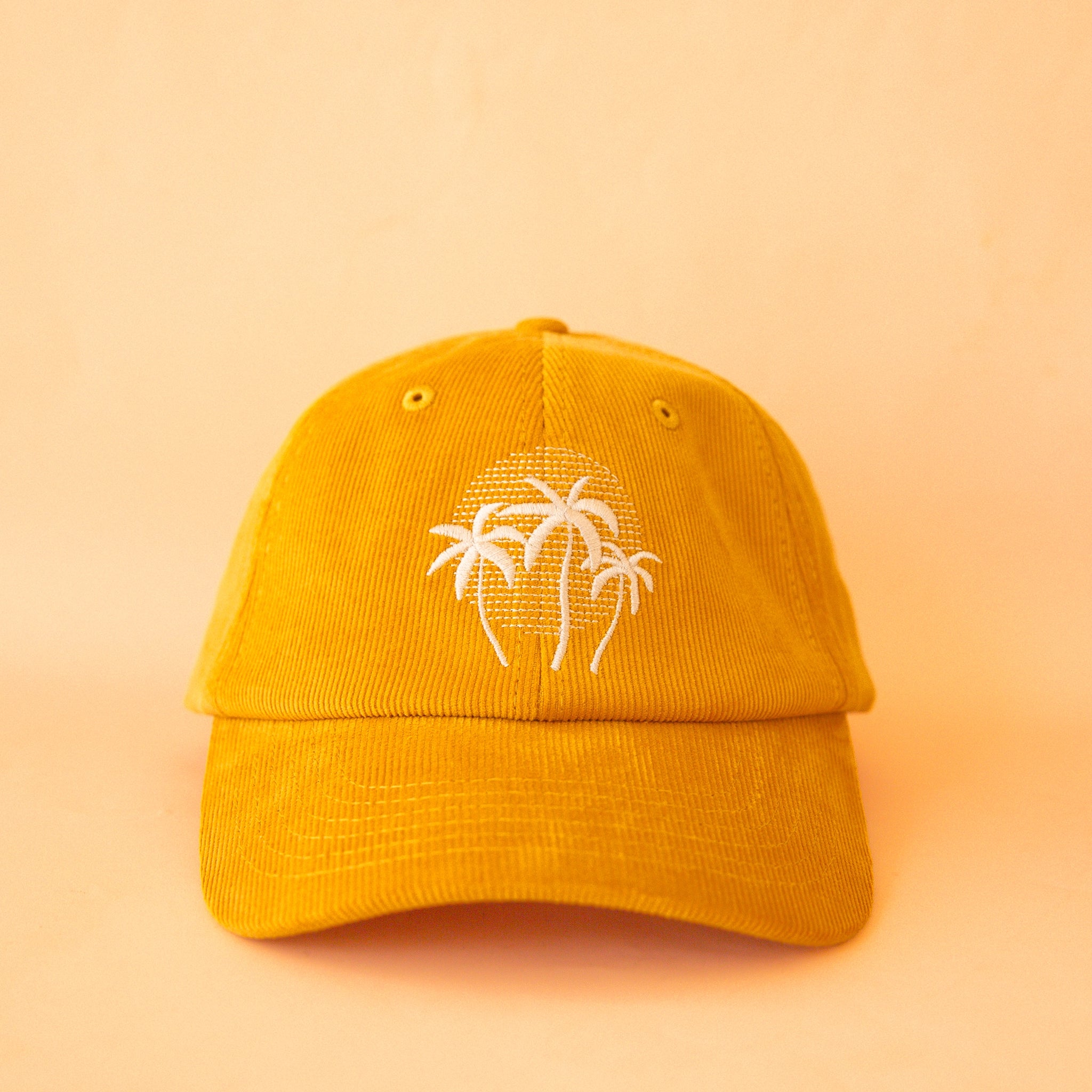 On a light yellow background is a golden yellow corduroy baseball hat with a white palm tree design in the center. 