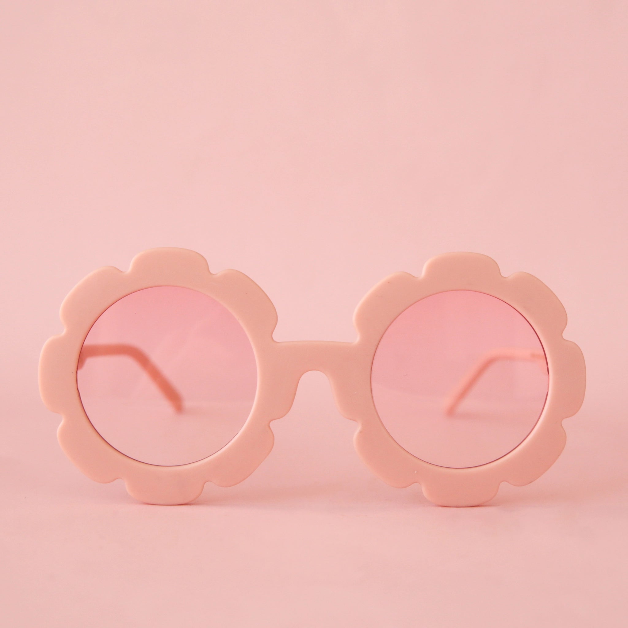 On a pink background is a light pink pair of flower shaped sunglasses with a light pink lens. 