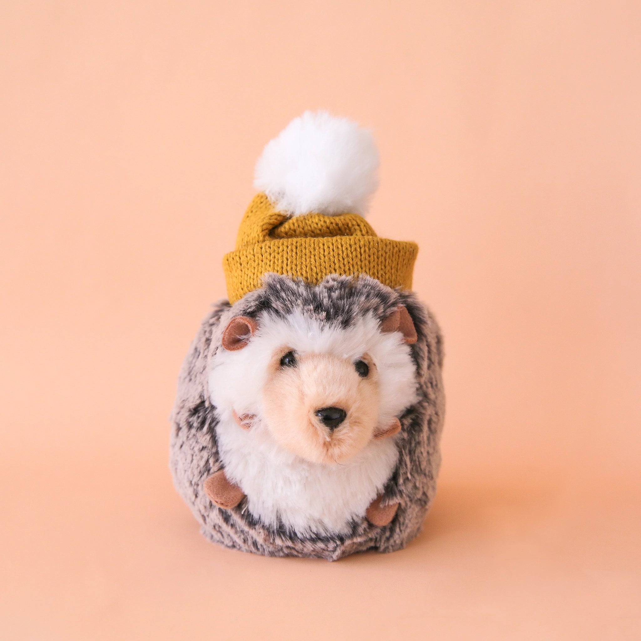 On a peachy background is a grey and white hedgehog stuffed animal with a mustard yellow knit beanie on its head with a white pom pom.