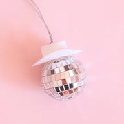 On a pink background is a disco ball car charm with a white cowboy hat and a string for hanging.