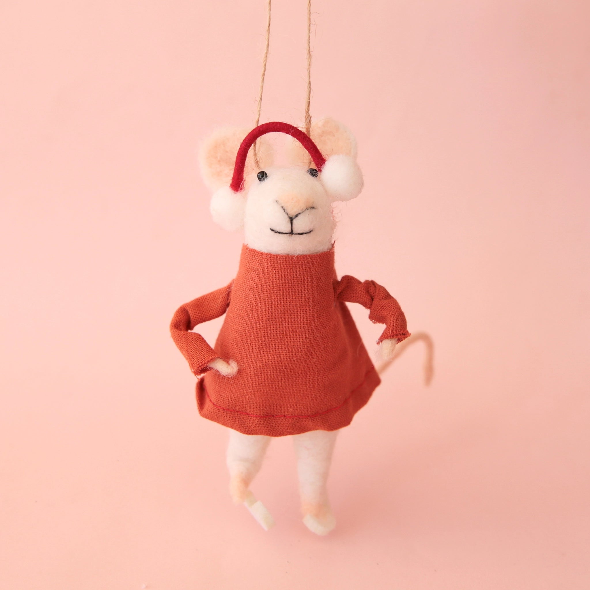 On a pink background is a white felt mouse ornament wearing a red dress and earmuffs and ice skates. 