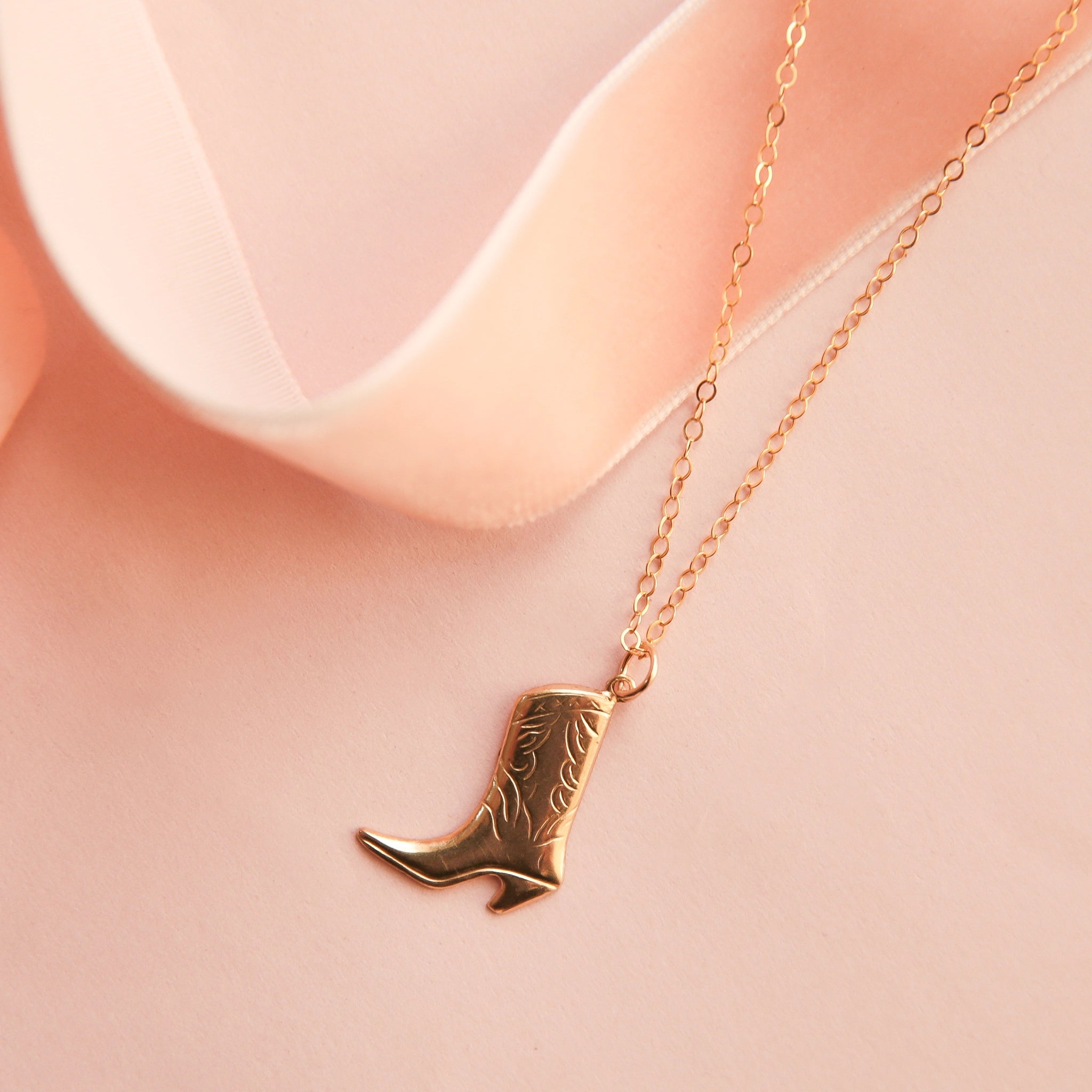 On a pink background is a gold chain necklace with a cowgirl boot pendant. 