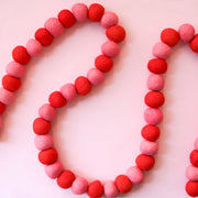 On a pink background is a felt ball garland made of pink and red alternating felt balls. 