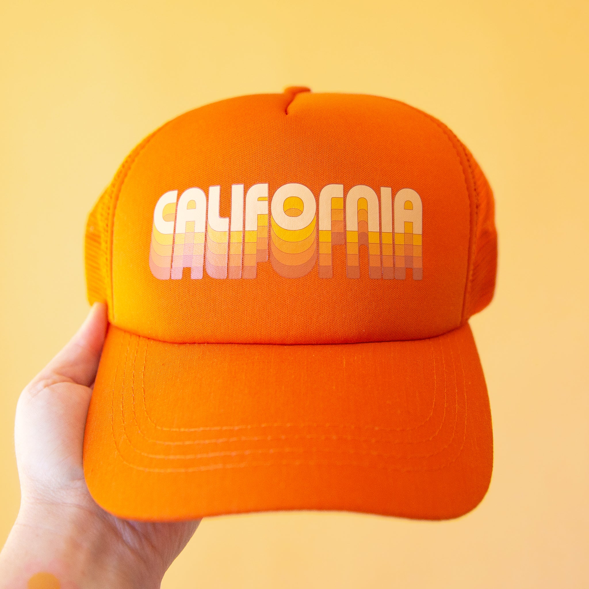 On a yellow background is an orange trucker hat with yellow text across the front that reads, &quot;California&quot;. 