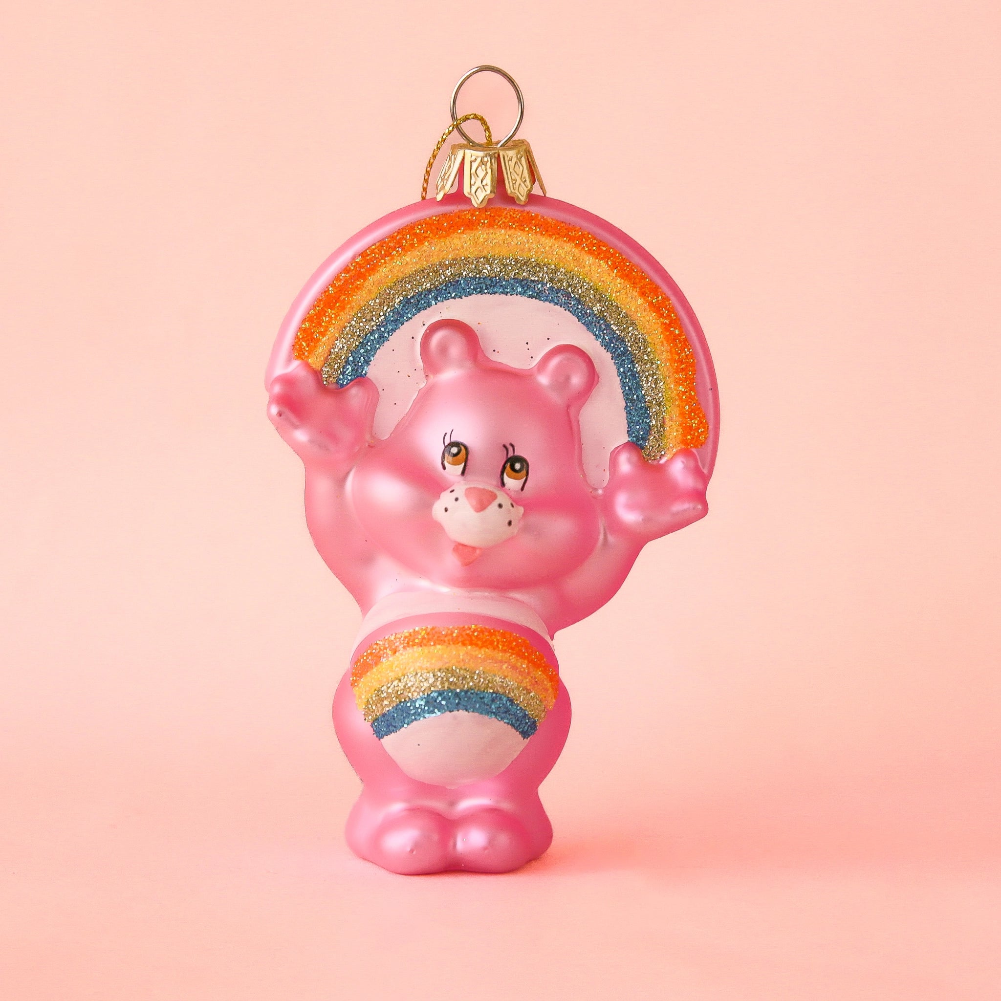 On a peachy background is a pink glass bear shaped ornament with a rainbow on its stomach and arched over its head. 