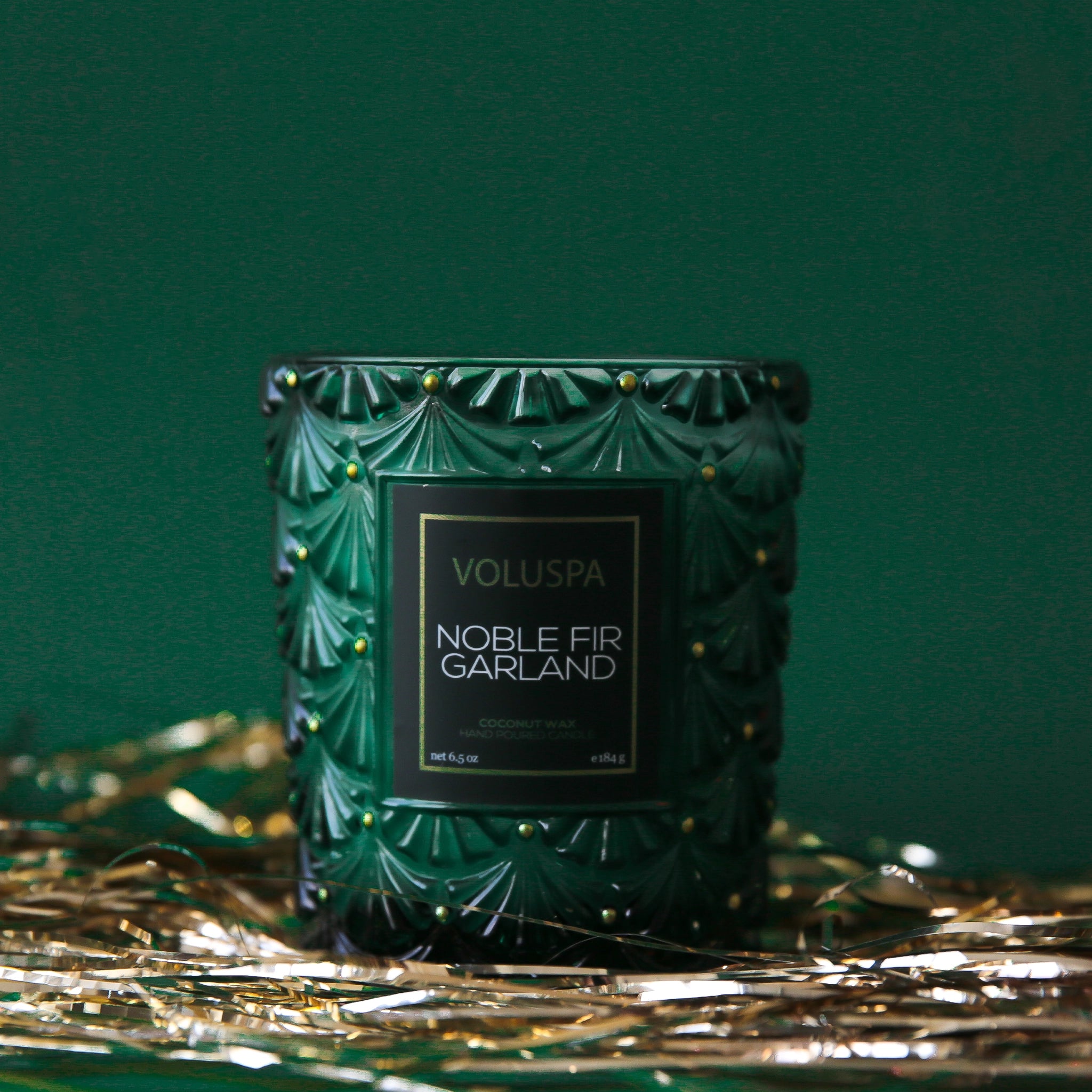 On a dark green background is a green glass candle with a label that reads, "Noble Fir Garland".