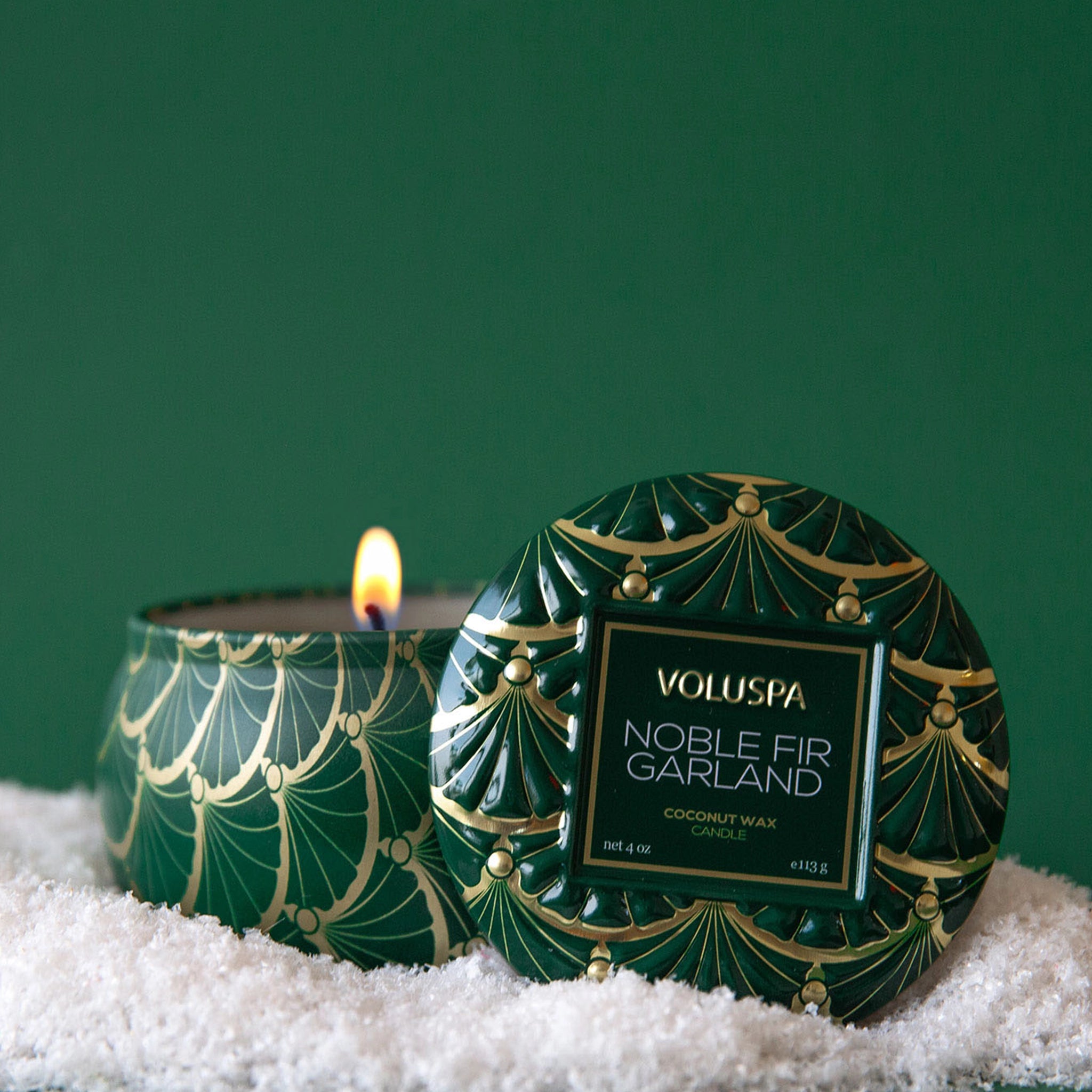 On a green background is a green metal mini tin candle with a gold and green design and a label on the lid that reads, "Voluspa Noble Fir Garland".