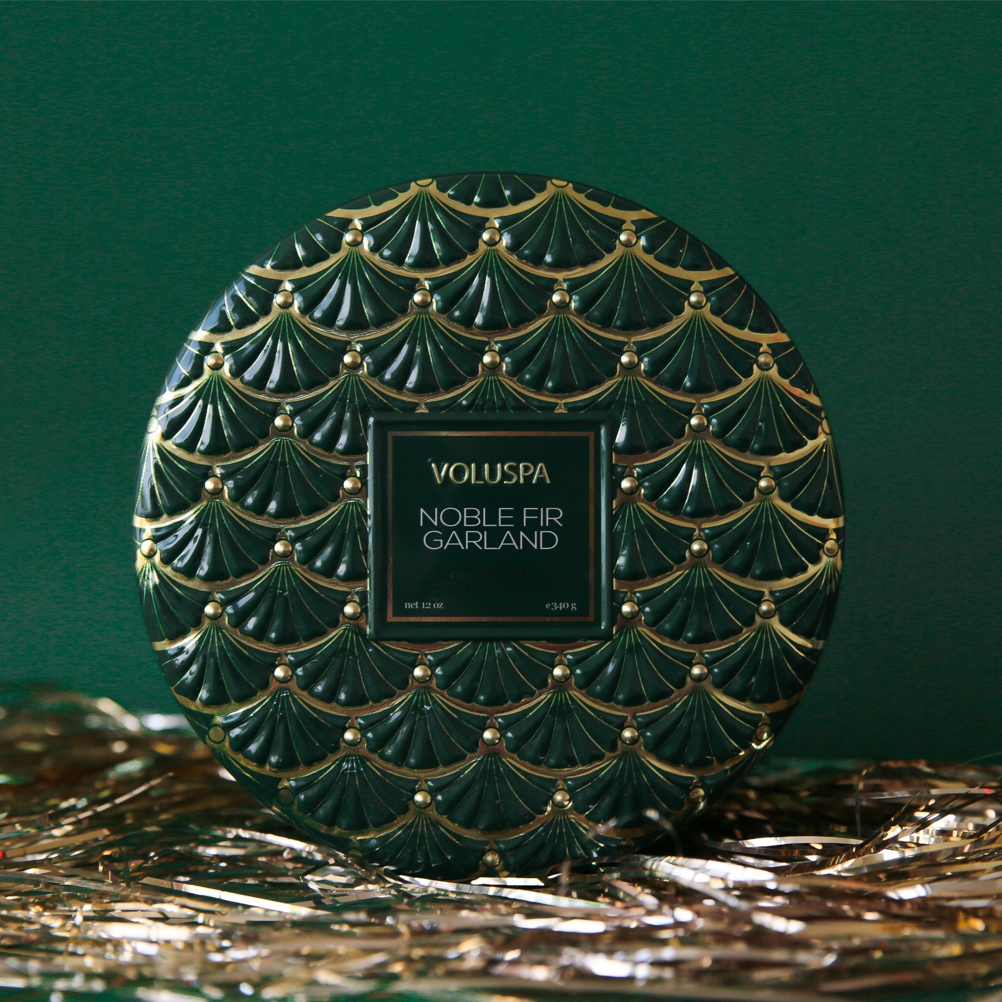 On a green background is a green 3 wick tin candle with a gold design and a matching lid.