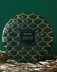 On a green background is a green 3 wick tin candle with a gold design and a matching lid.