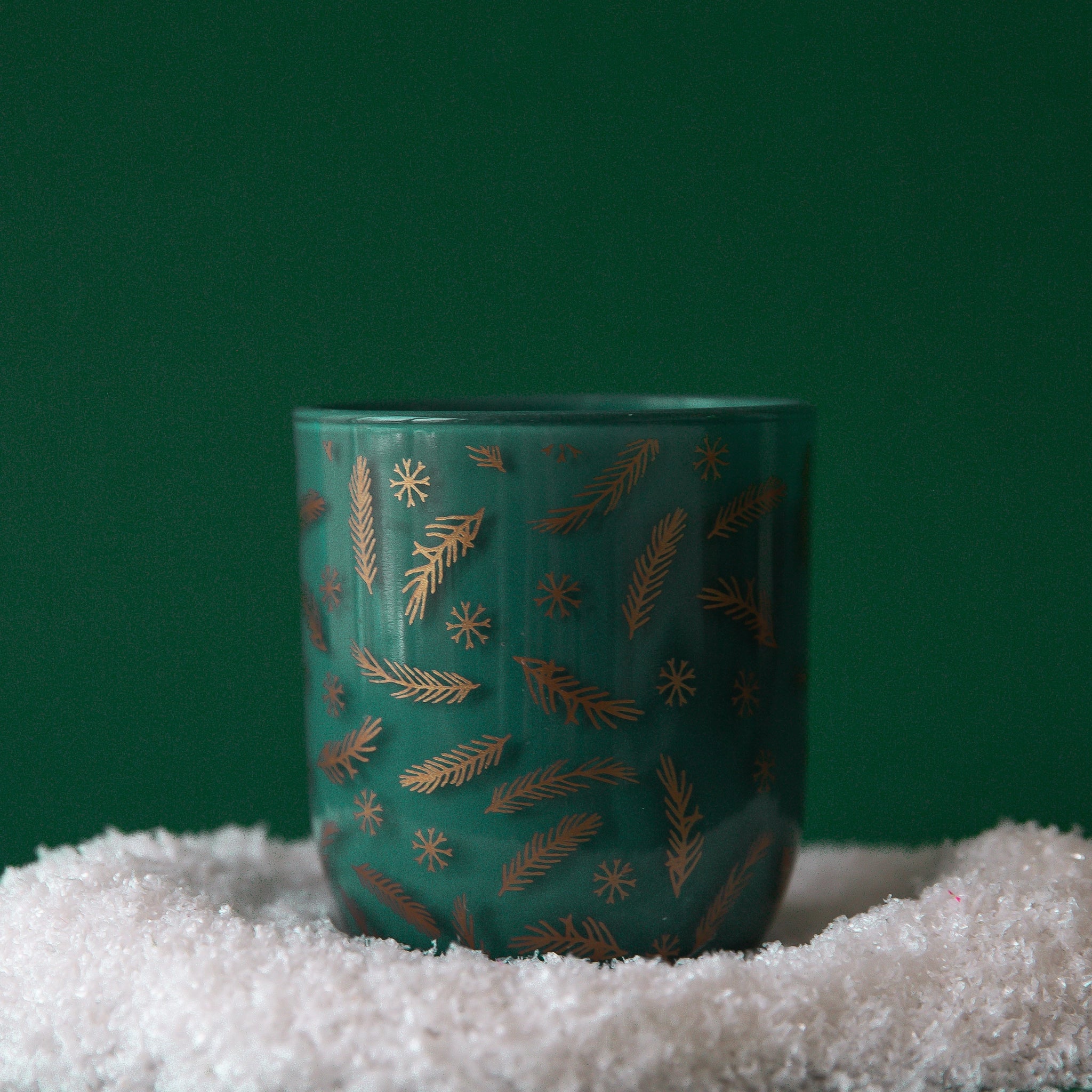 On a green background is a green and gold glass candle with a snowflake and pine needle pattern.