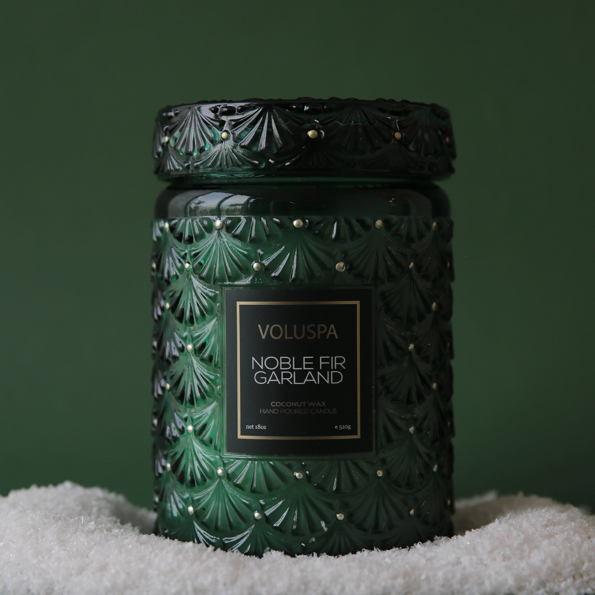 On a green background is a dark green decorative glass jar candle with gold dot details and a label in the center that reads, &quot;Noble Fir Garland&quot;.