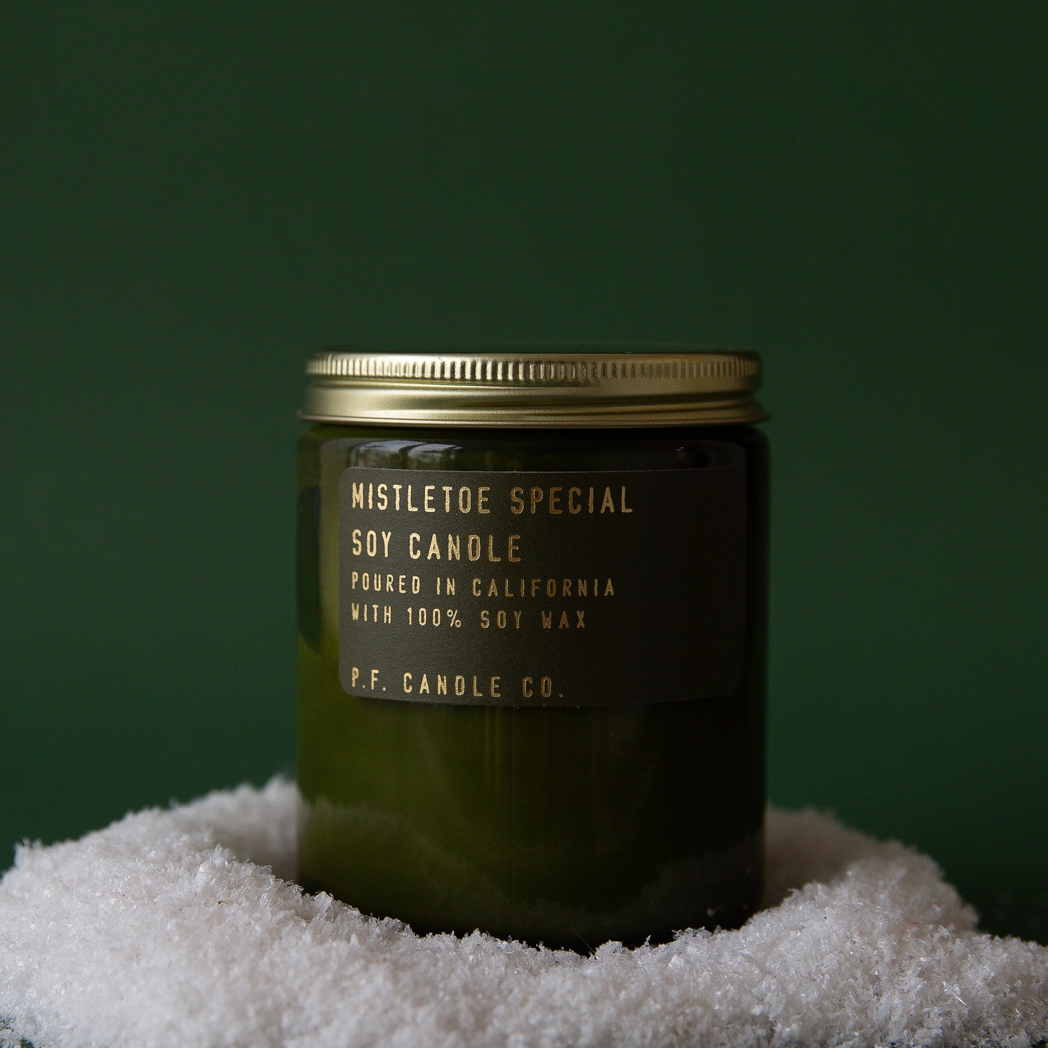 On a dark green background is a green glass jarred candle with a gold lid and gold lettering that reads, &quot;Mistletoe Special Soy Candle&quot;.