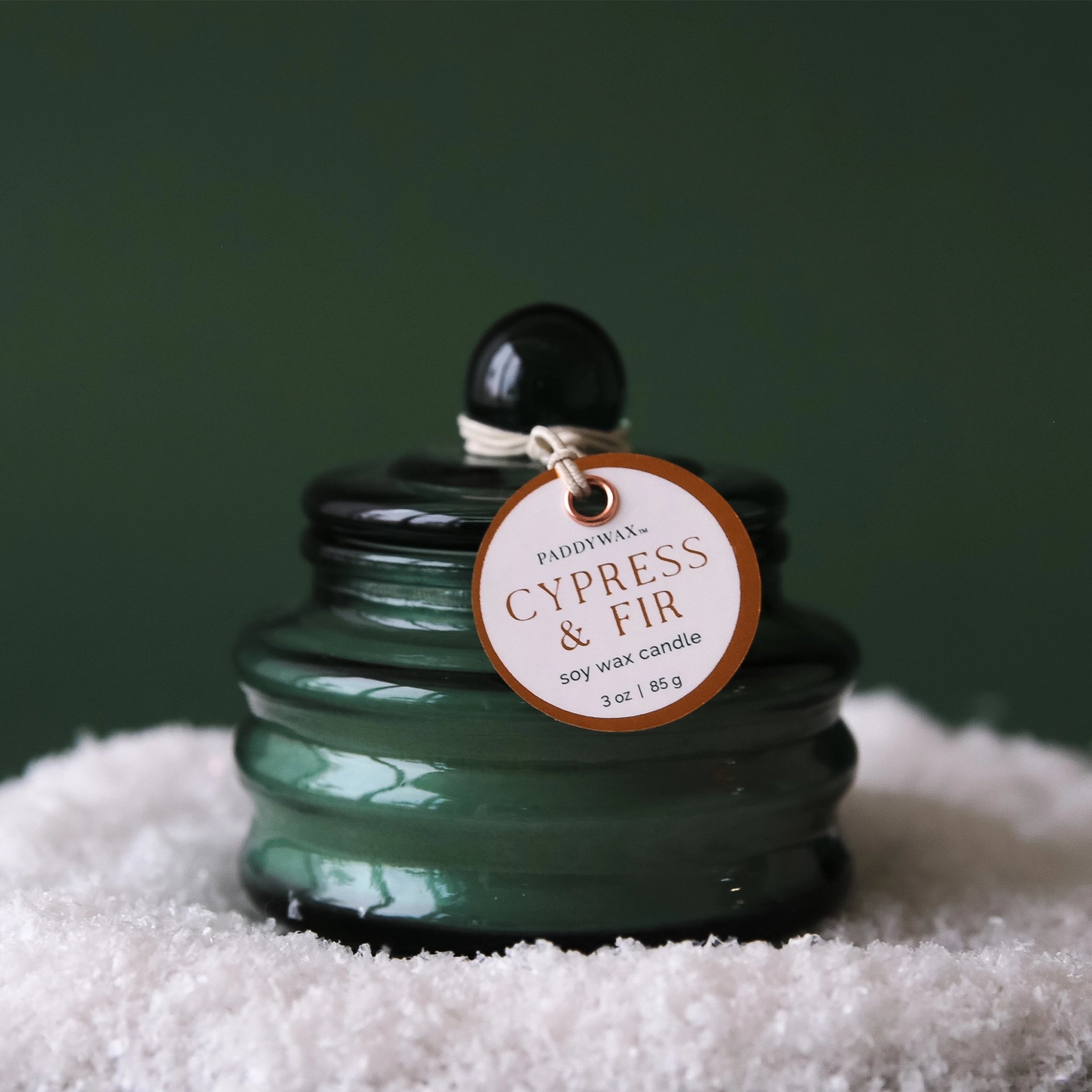 On a peachy background is a dark green glass candle with a coordinating lid and a round label tag that reads, &quot;Cypress &amp; Fir Soy Wax Candle&quot;.