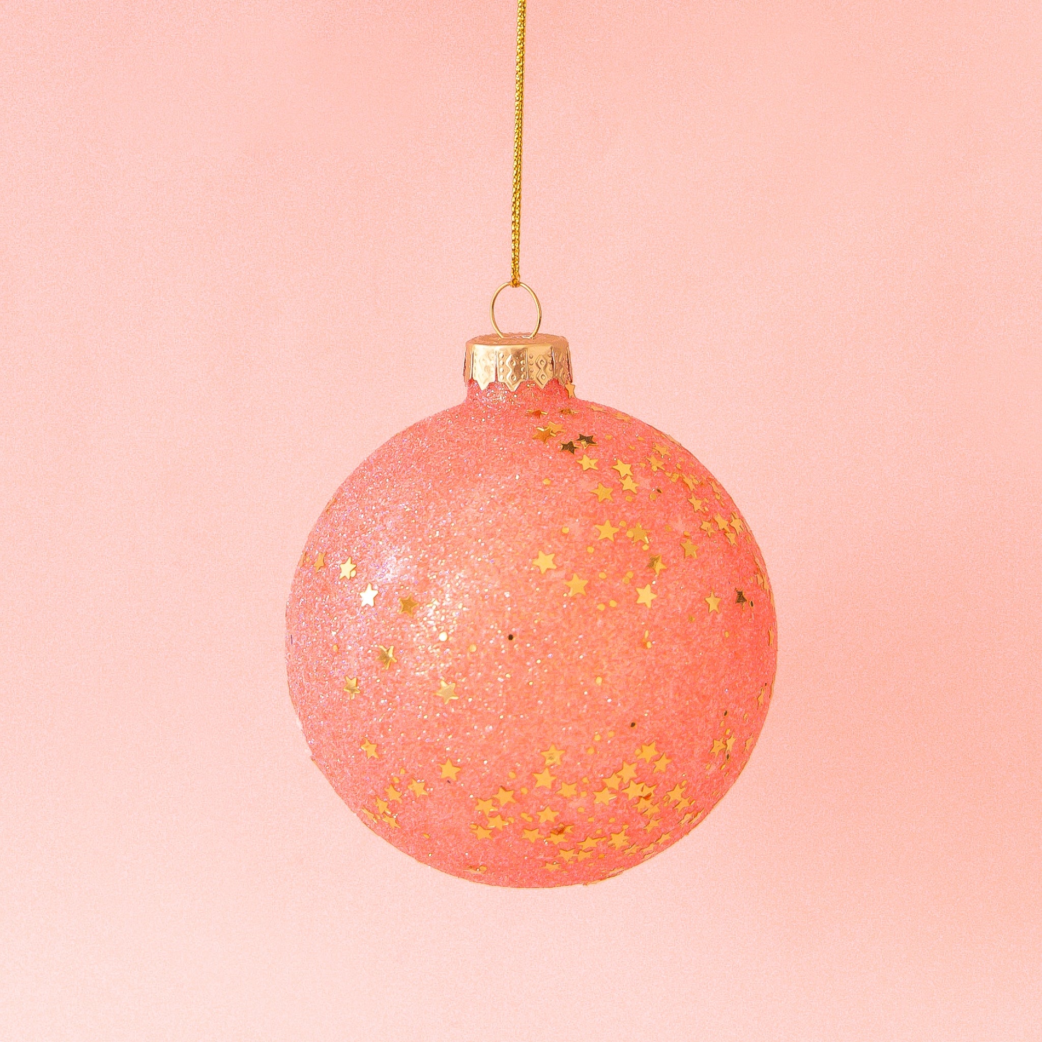 On a pink background is a watermelon colored glass ball ornament with a gold sparkle and star detail.