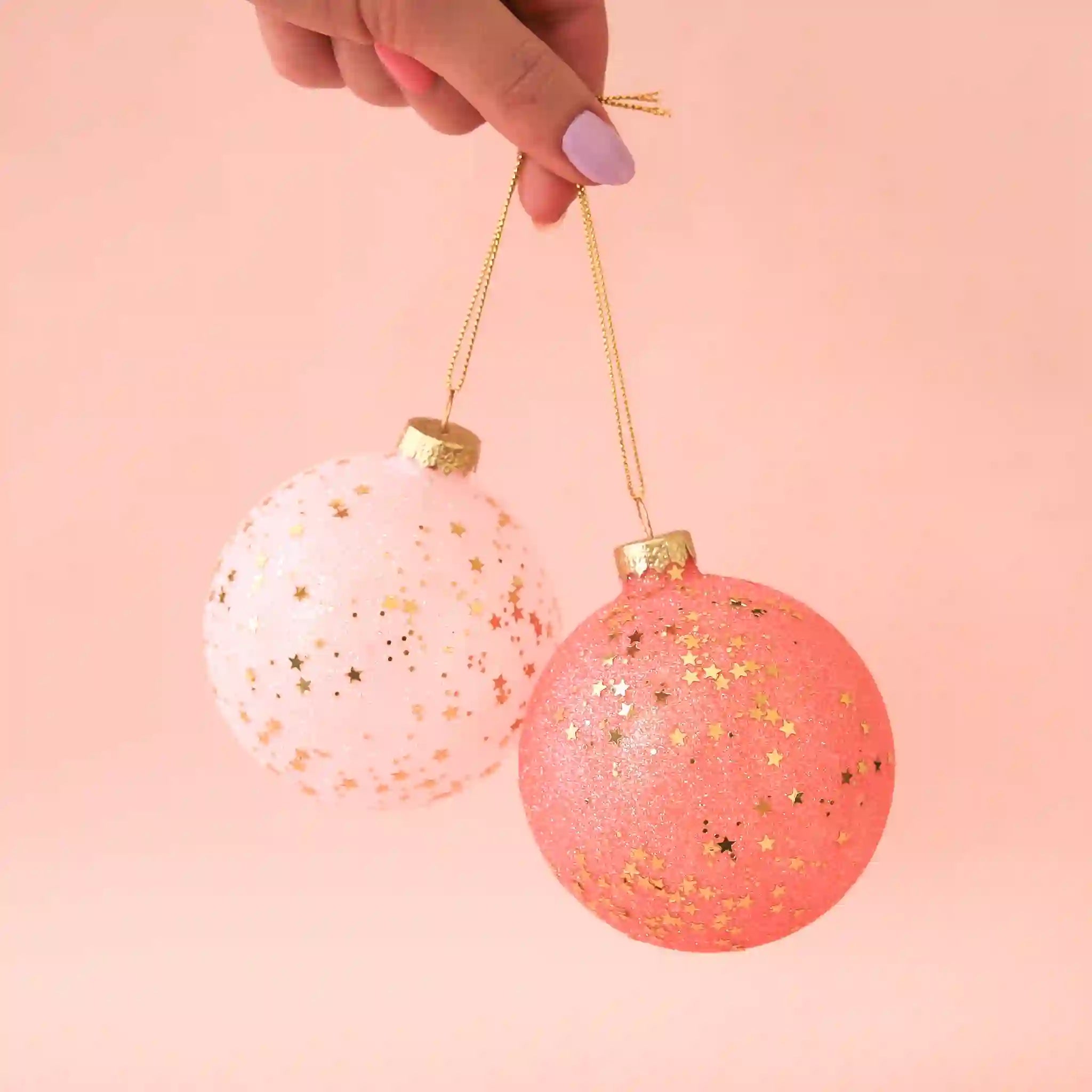 On a peachy background is two different tones of pink ball ornaments with gold glitter and stars. 