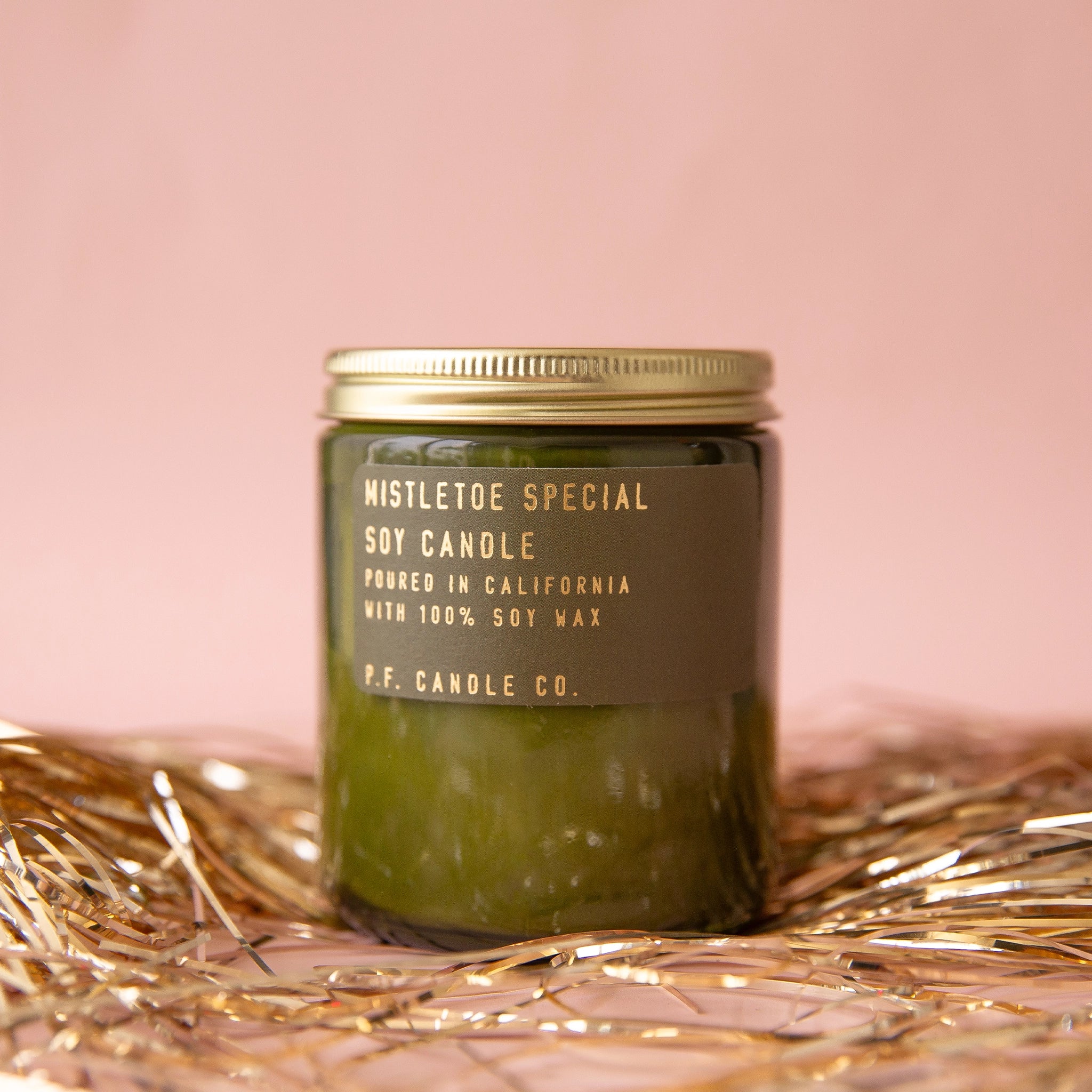 On a pink background is a green glass jarred candle with a gold lid and gold lettering that reads, "Mistletoe Special Soy Candle".