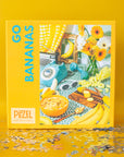 On a yellow background is a red and blue puzzle box ghat reads, "Go Bananas" with a photo on the side of what the puzzle looks like finished which is a retro table scape with bananas a vintage touch dial phone yellow flowers and white oval sunglasses. 