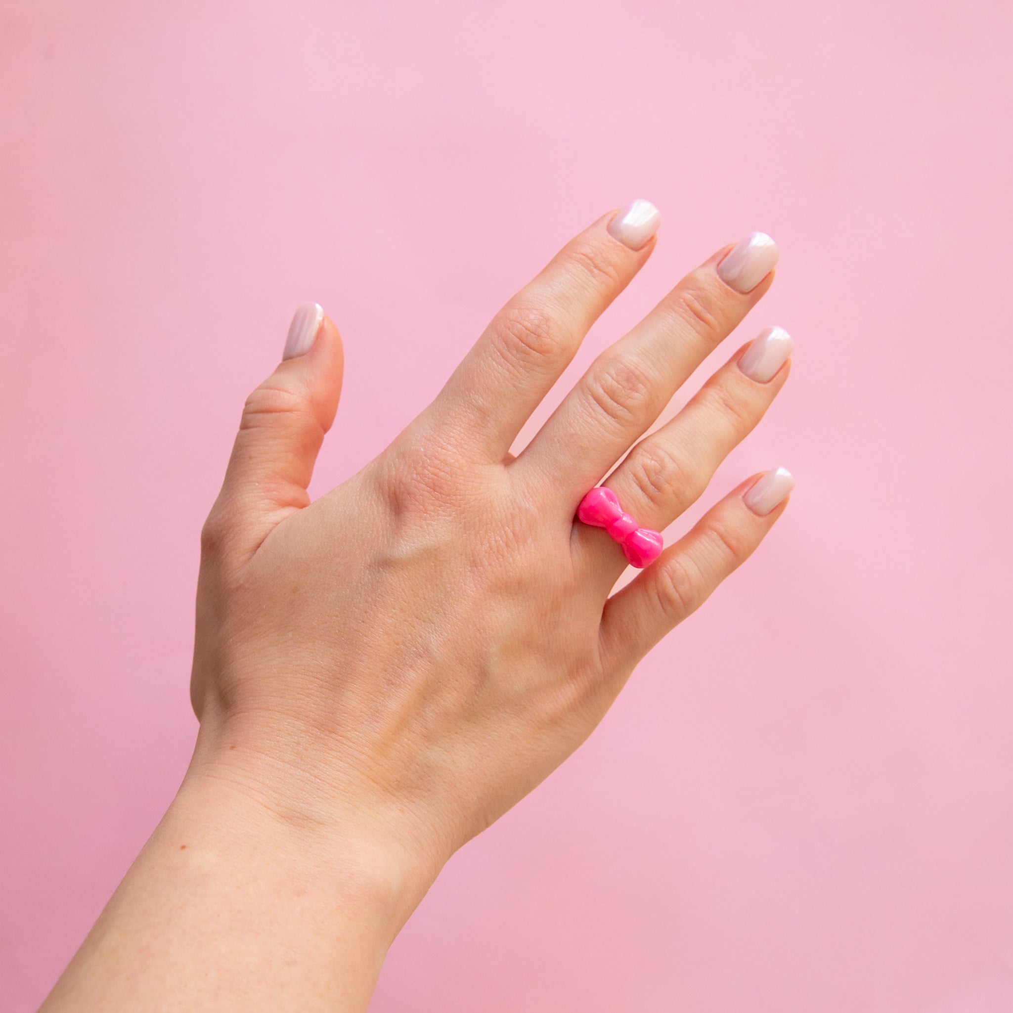 A hand wearing a cute acrylic bow shaped ring in pink with a pink background.