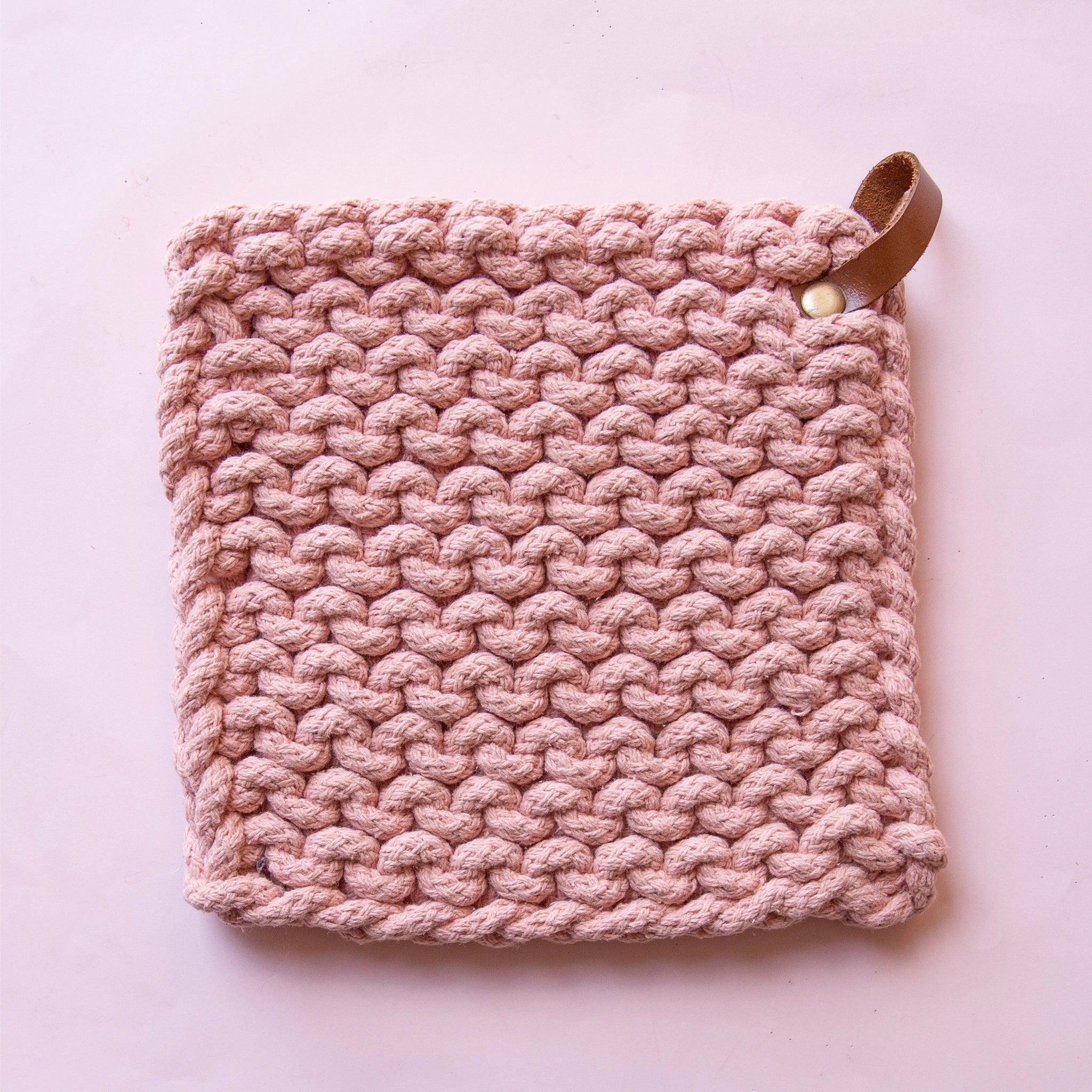On a pink background is a rose colored crocheted pot holder with a leather loop for hanging in the top right corner. 