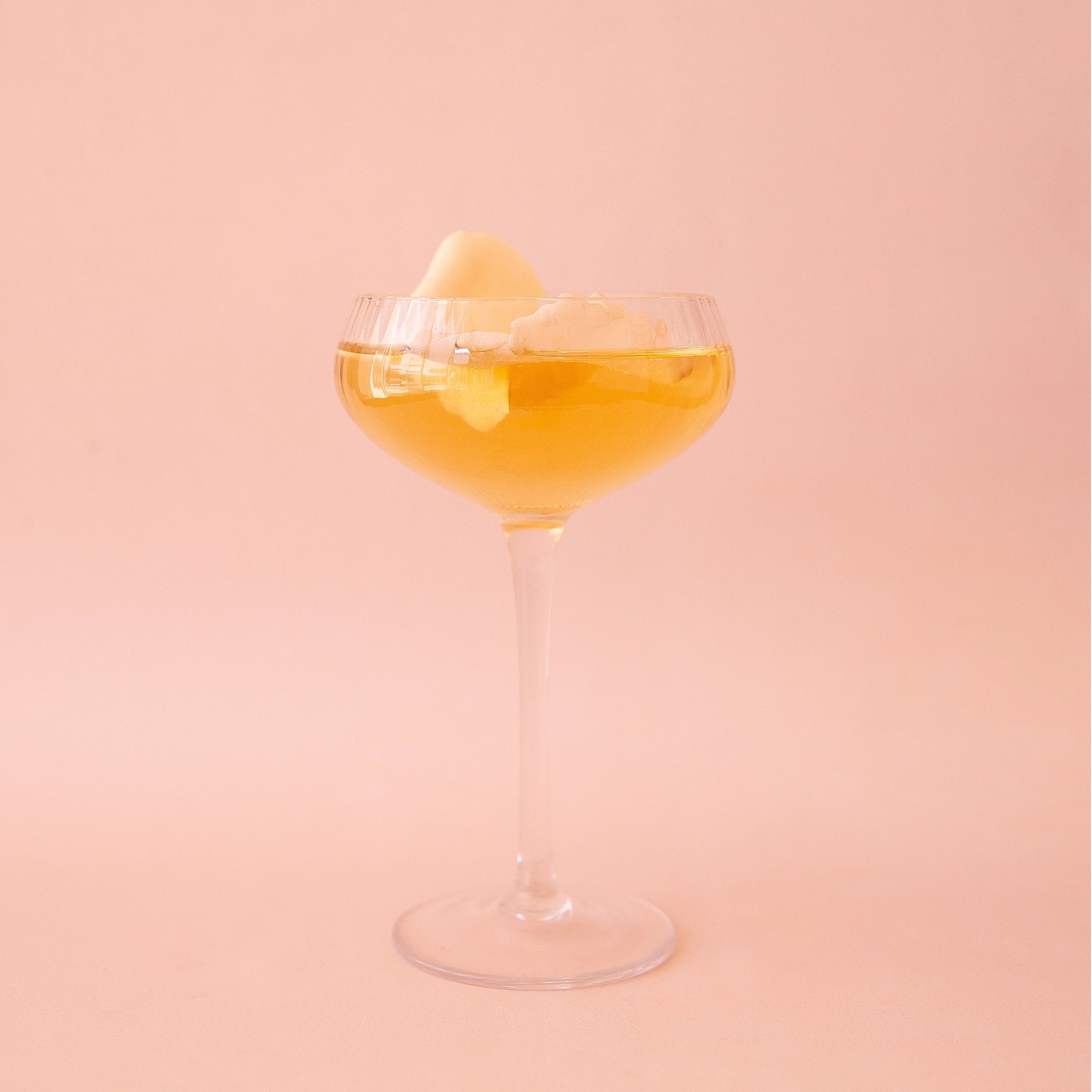 On a pink background is a thin clear coupe glass with a bevelled texture and a yellow liquid inside. 