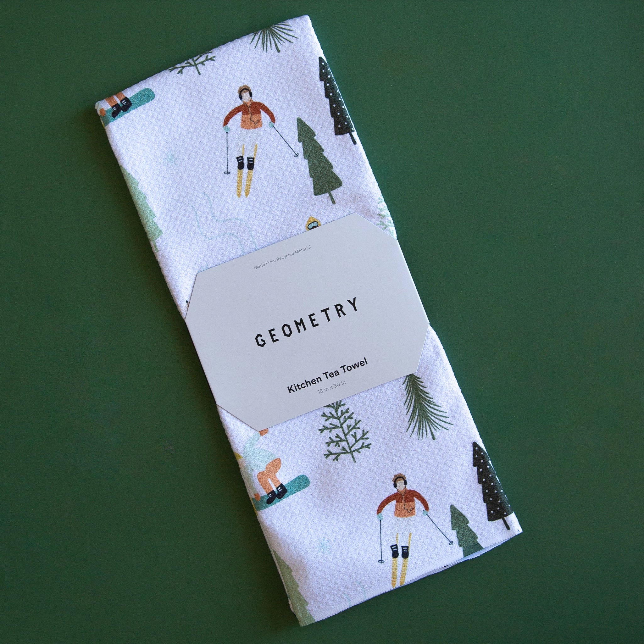 On a green background is a folded white waffle knit kitchen towel with a ski and snowboard pattern. 