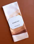 On a brown background is a white and warm toned waffle knit tea towel. 