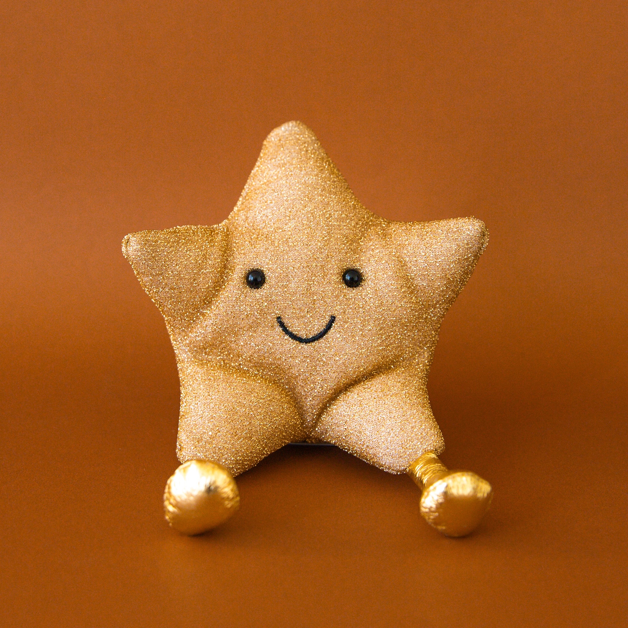 On a burnt orange background is a gold star stuffed toy with a smiling face and gold legs. 