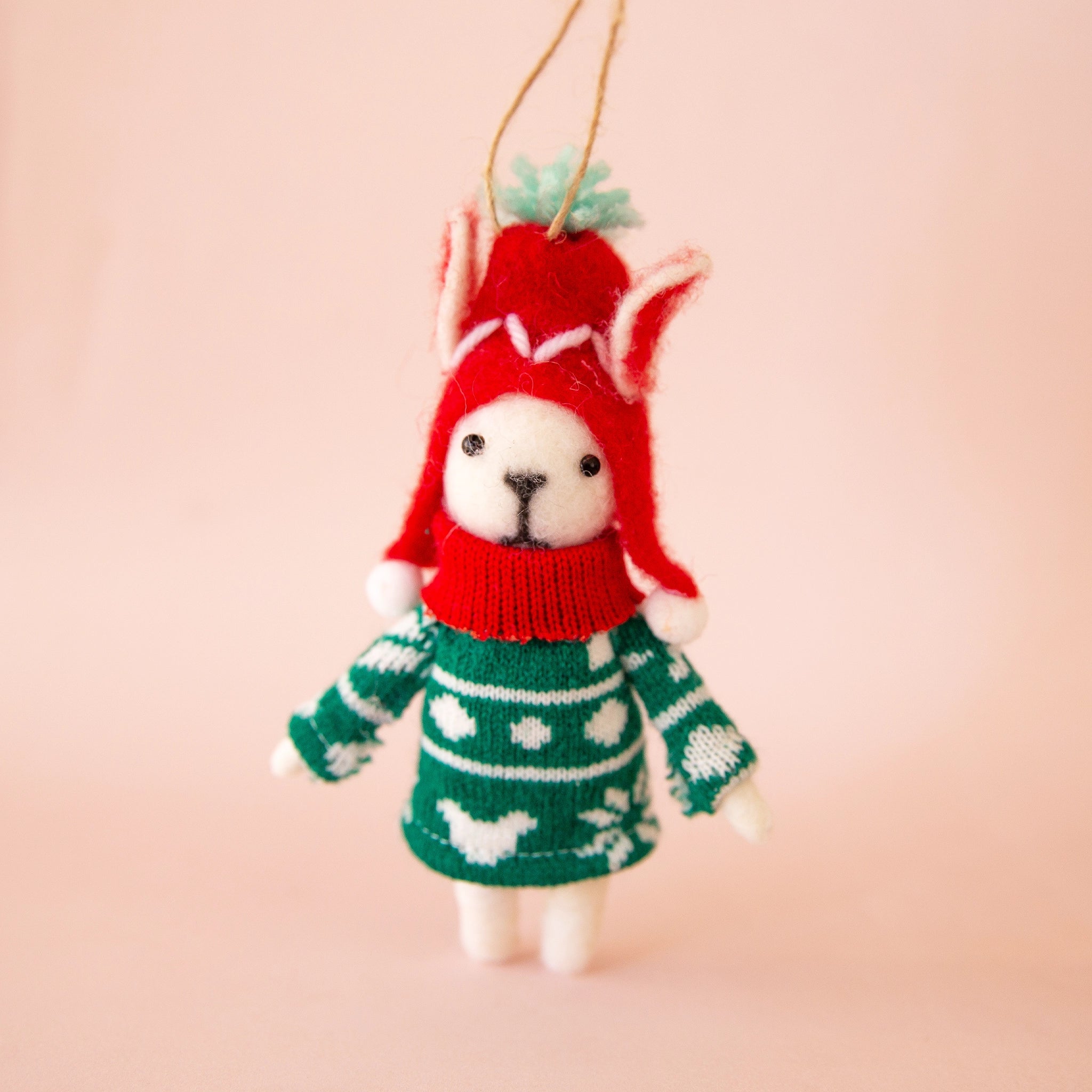 On a peachy background is a white felt dog ornament with a green sweater and red hat on. 