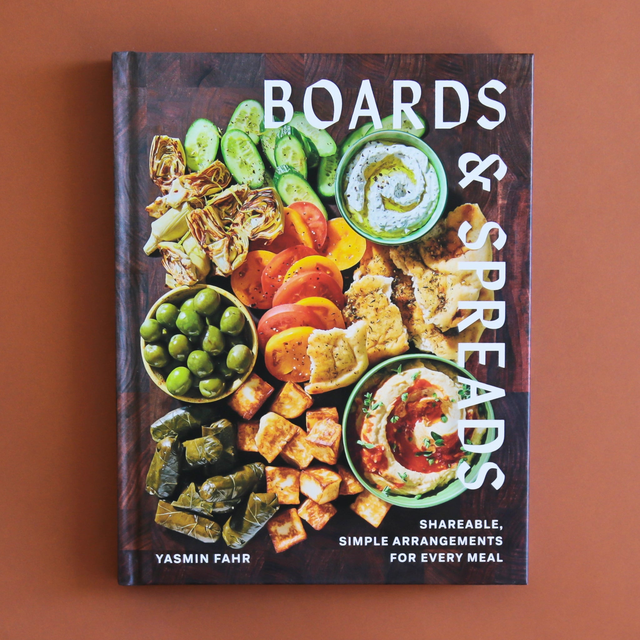 A book cover mimicking the look of a cutting board with a variety of charcuterie style foods, like olives, spreads, veggies and more.