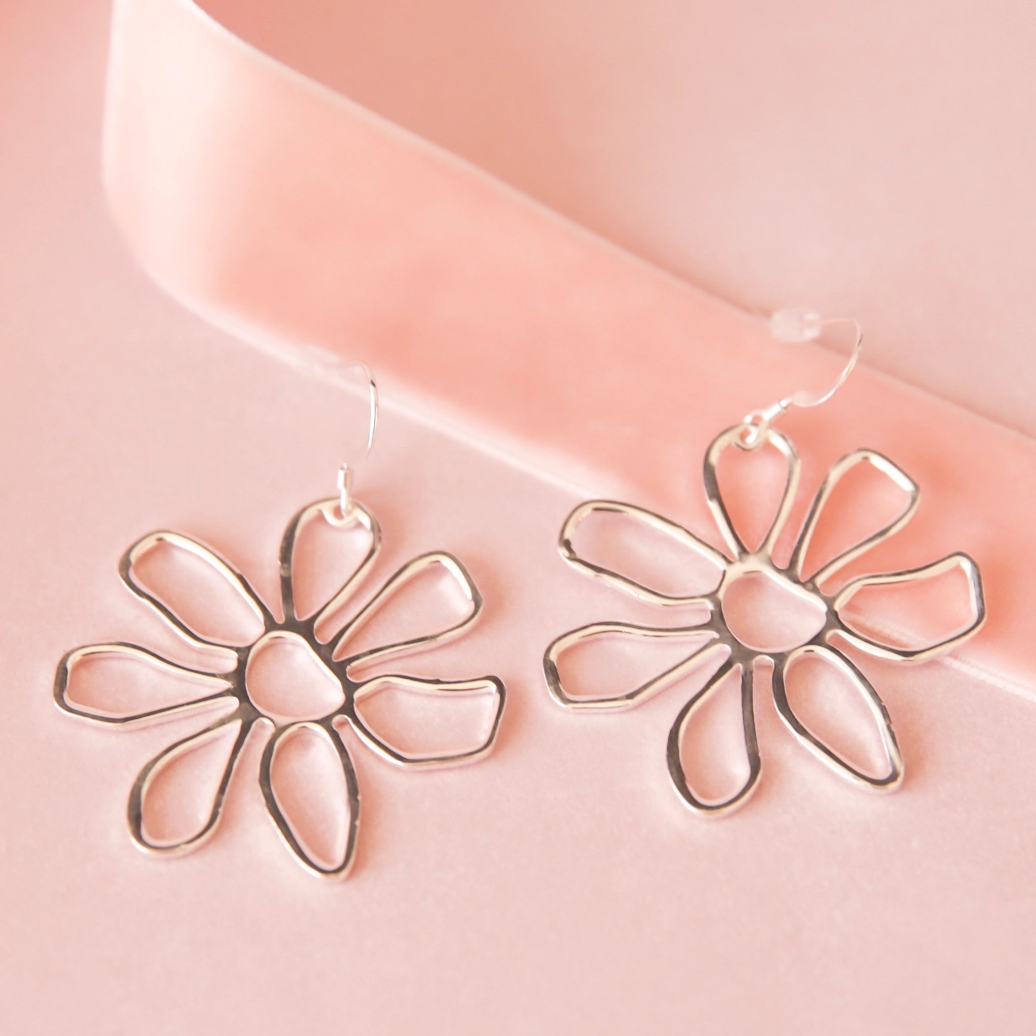 A pair of silver floral earrings lay on a pink background