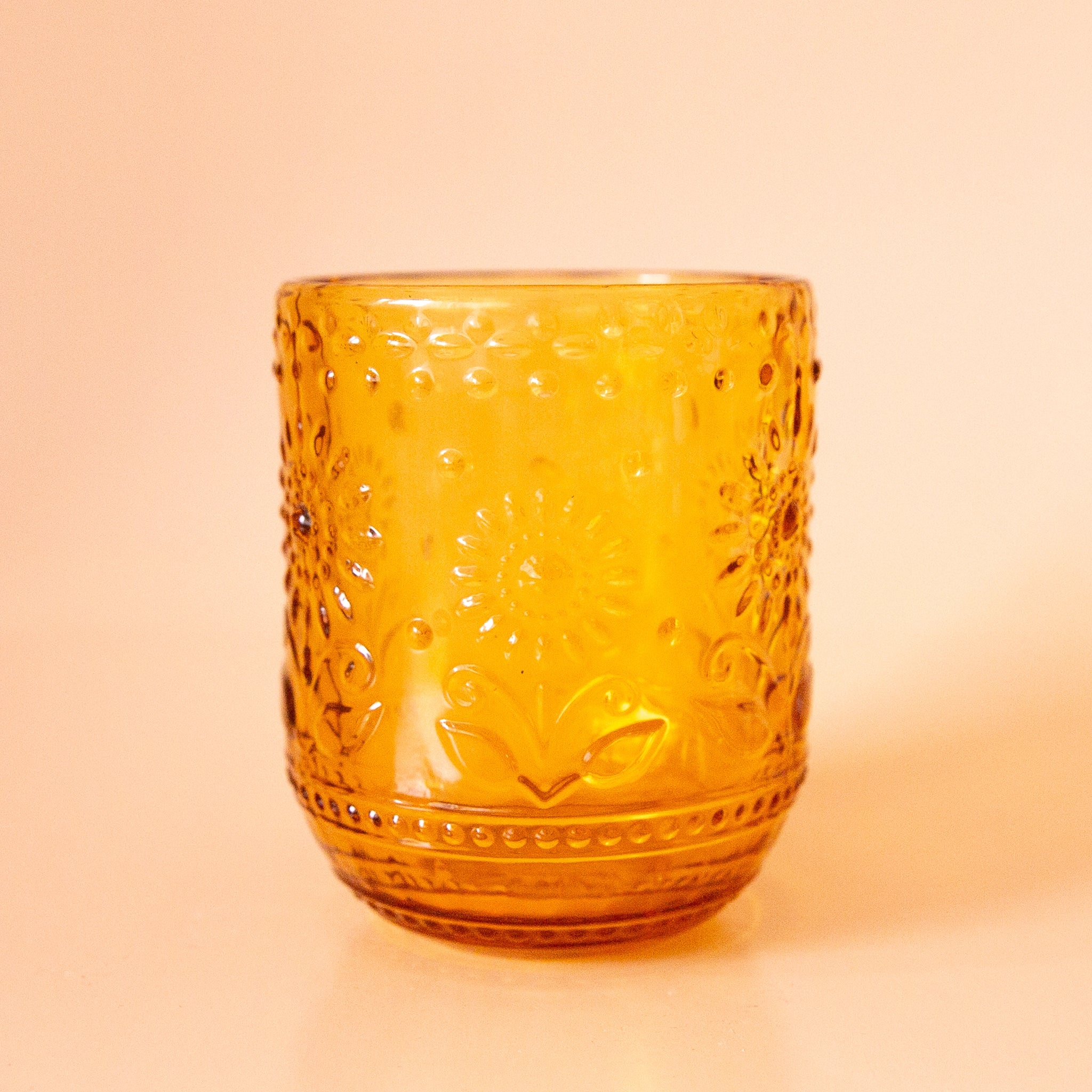 On a peachy background is an embossed yellow drinking glass with raised floral detailing.