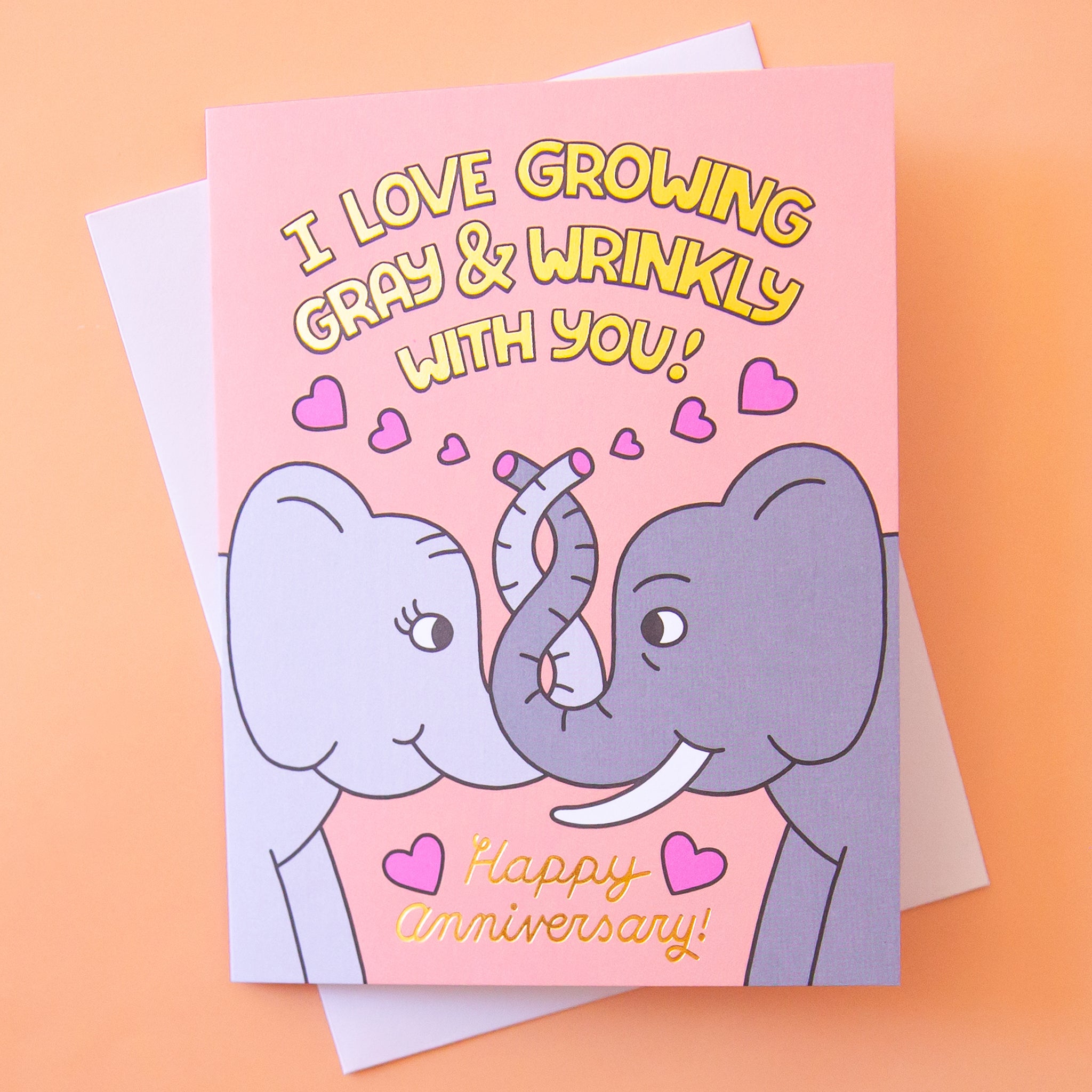 On a orange background is a card with a graphic of elephants intertwining trunks and text above and below that reads, "I Love Growing Gray & Wrinkly With You! Happy Anniversary". 