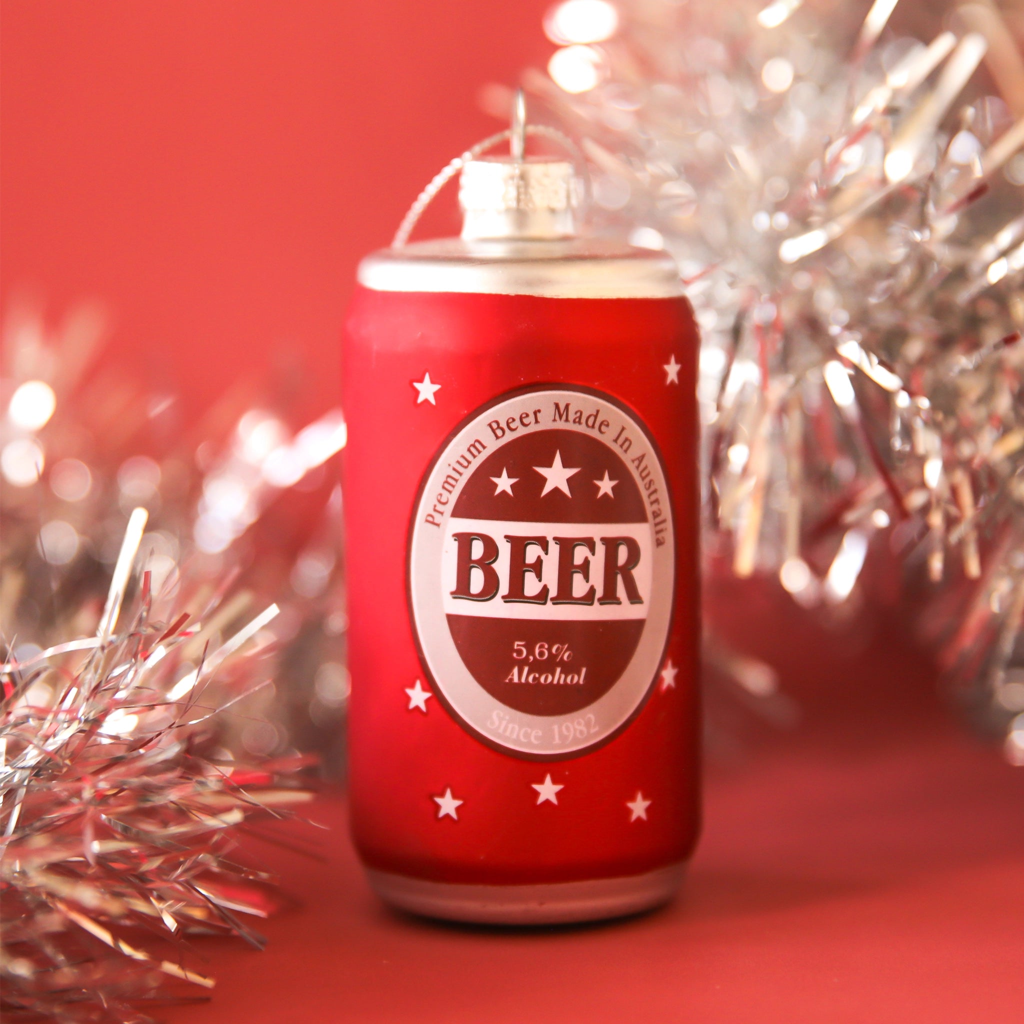 On a red tinsel background is a glass beer can shaped ornament. 