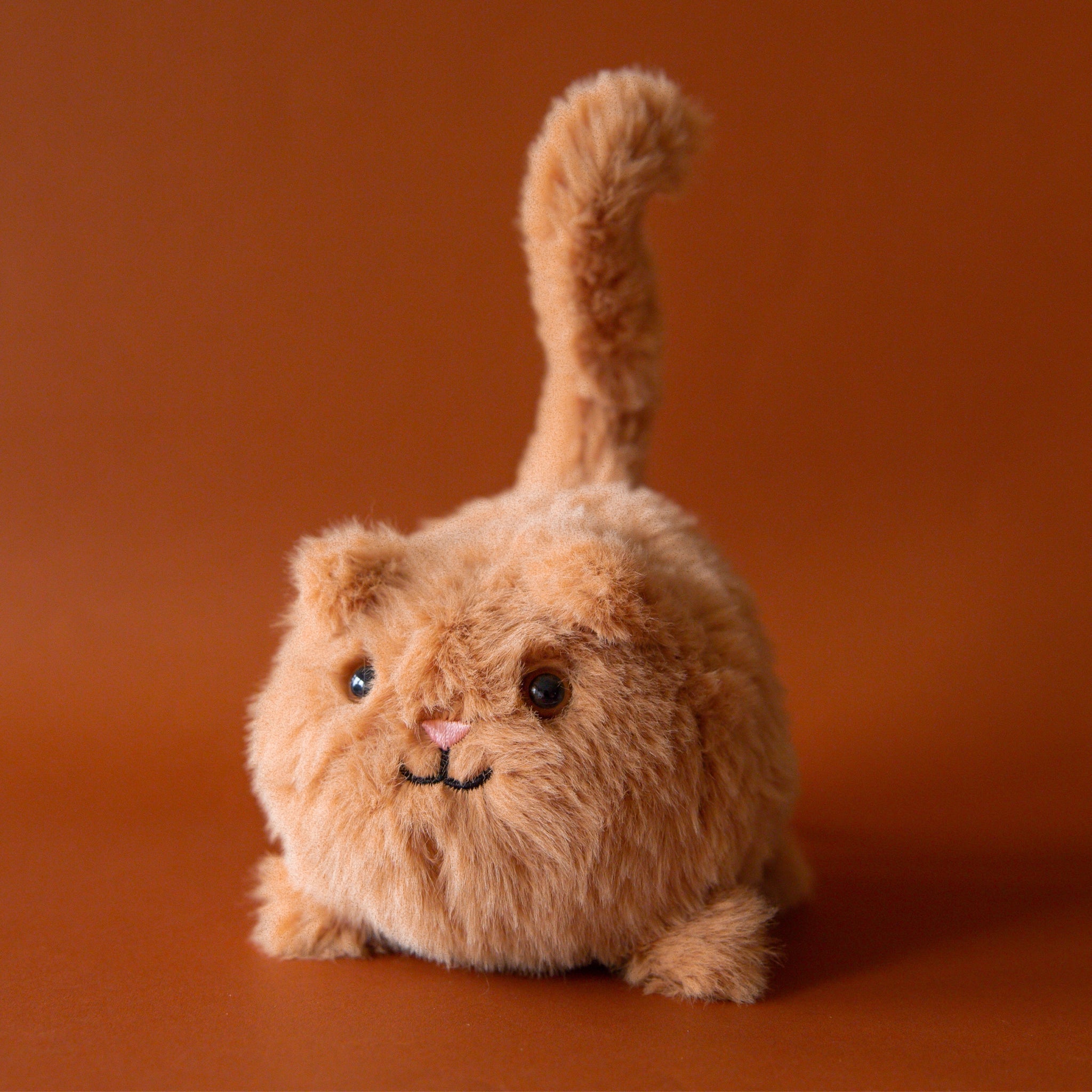 Soft stuffed animal in the shape of a round caramel colored cat with a question mark shaped tail