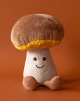 A soft suede stuffed animal in the shape of a mushroom with light brown cap with curly soft gold underneath, a tan colored stalk with smiley face and brown floppy legs.